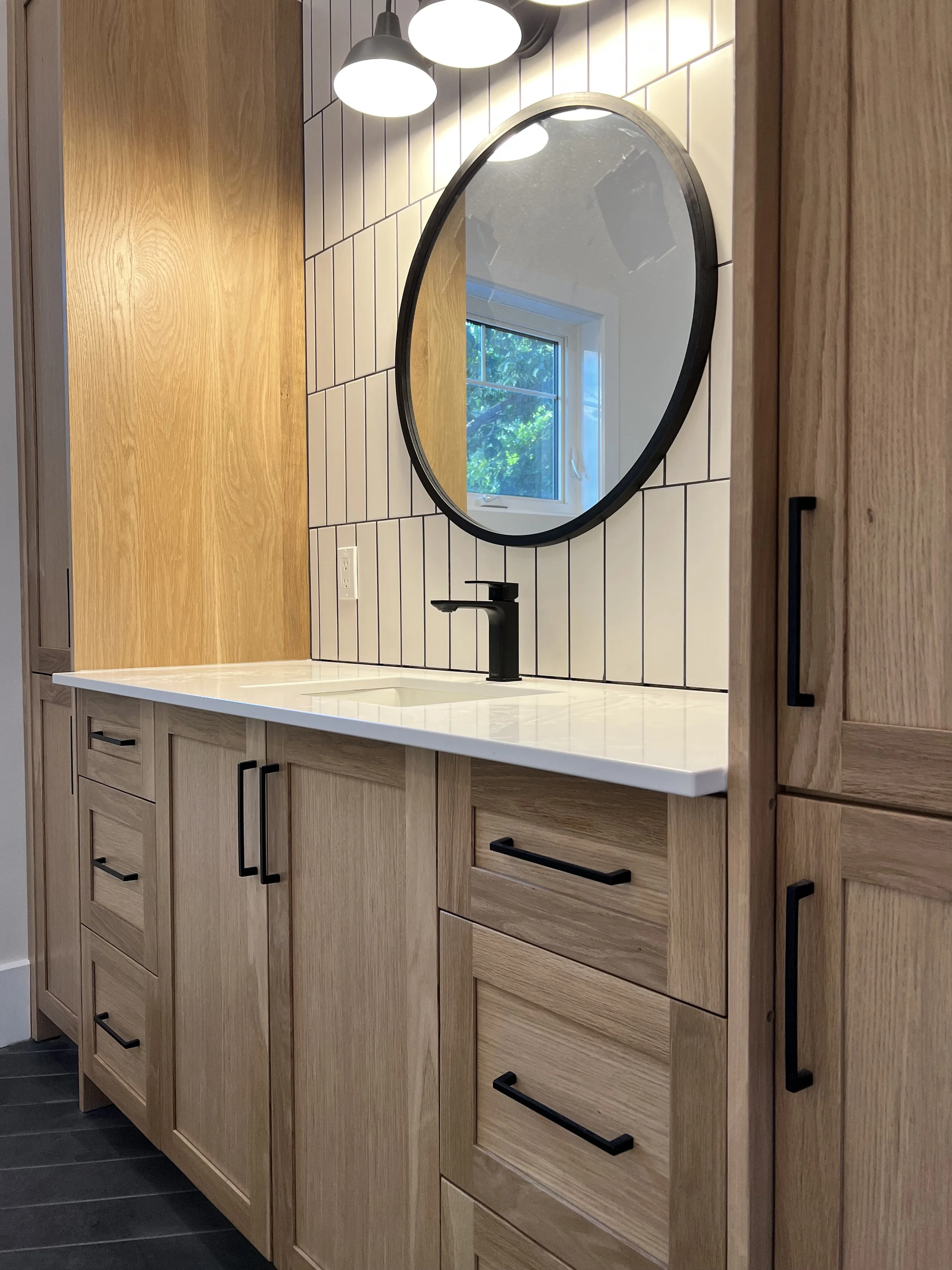 A modern bathroom vanity area with wooden cabinets, a white countertop, a black faucet, a large oval mirror, and a window reflecting greenery outside. The wall behind the mirror is tiled with white square tiles, and there is a black light fixture above the mirror.