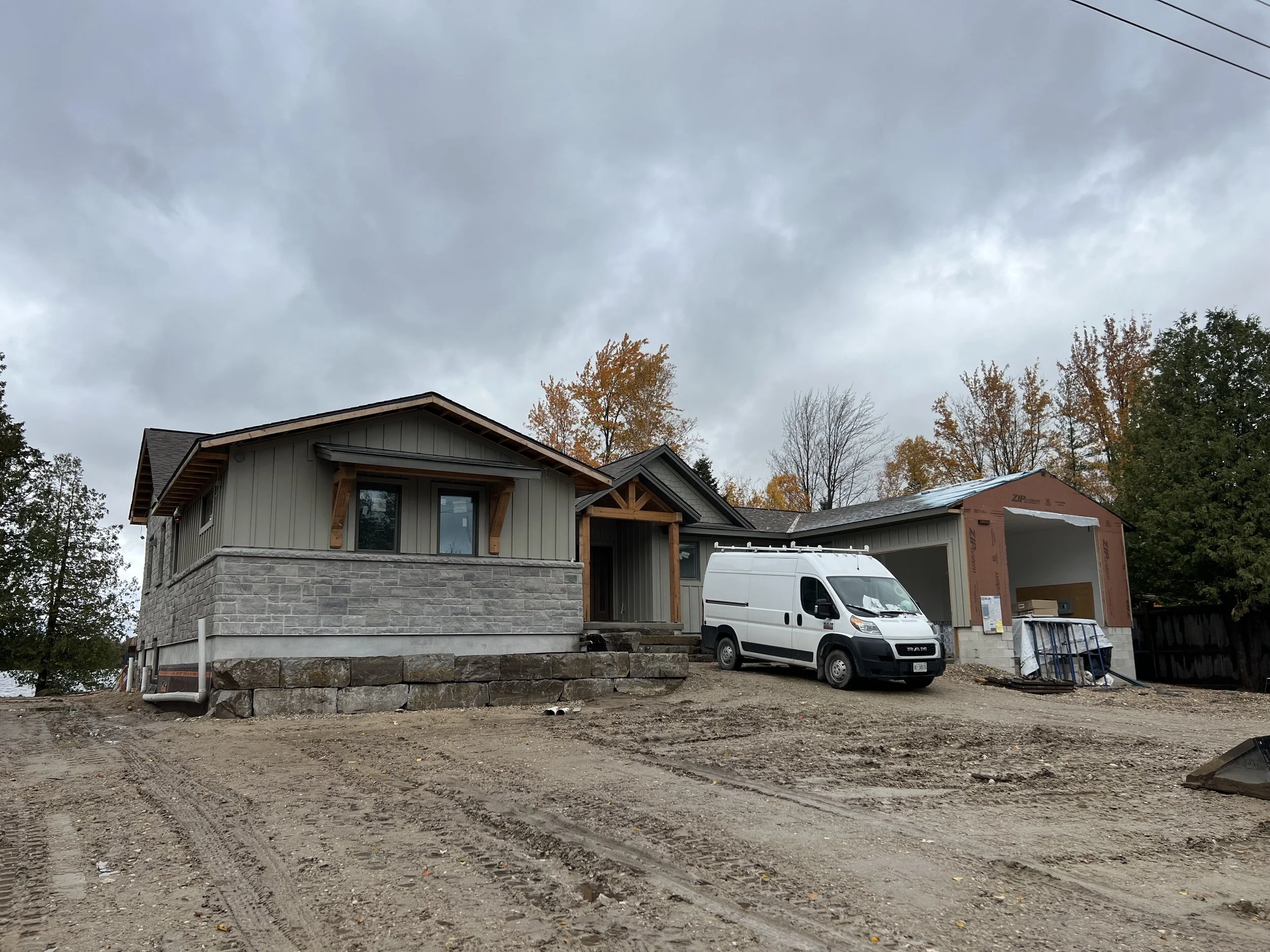 A house under construction with a gray exterior, a stone foundation, and a garage. A white van is parked in the driveway. The sky is overcast, and there are trees with autumn foliage in the background.
