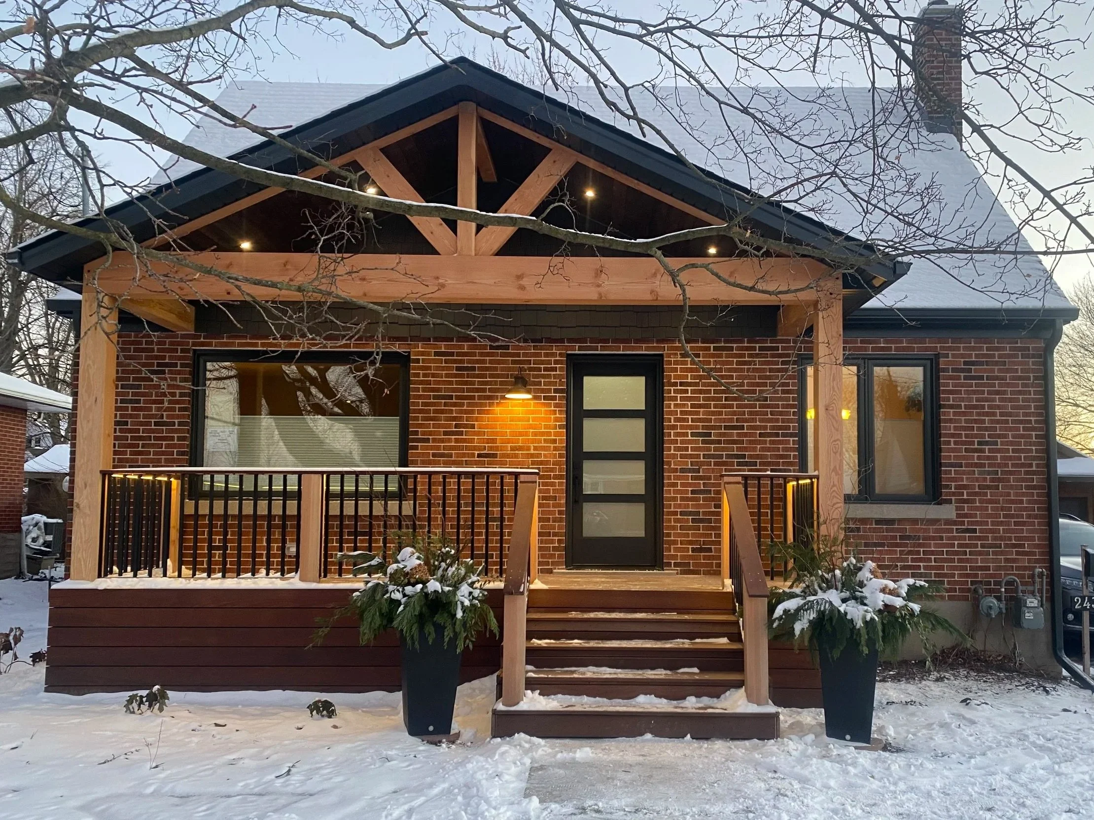 A brick house with a porch under construction, surrounded by snow, with a tree in the foreground and a chimney on the roof.