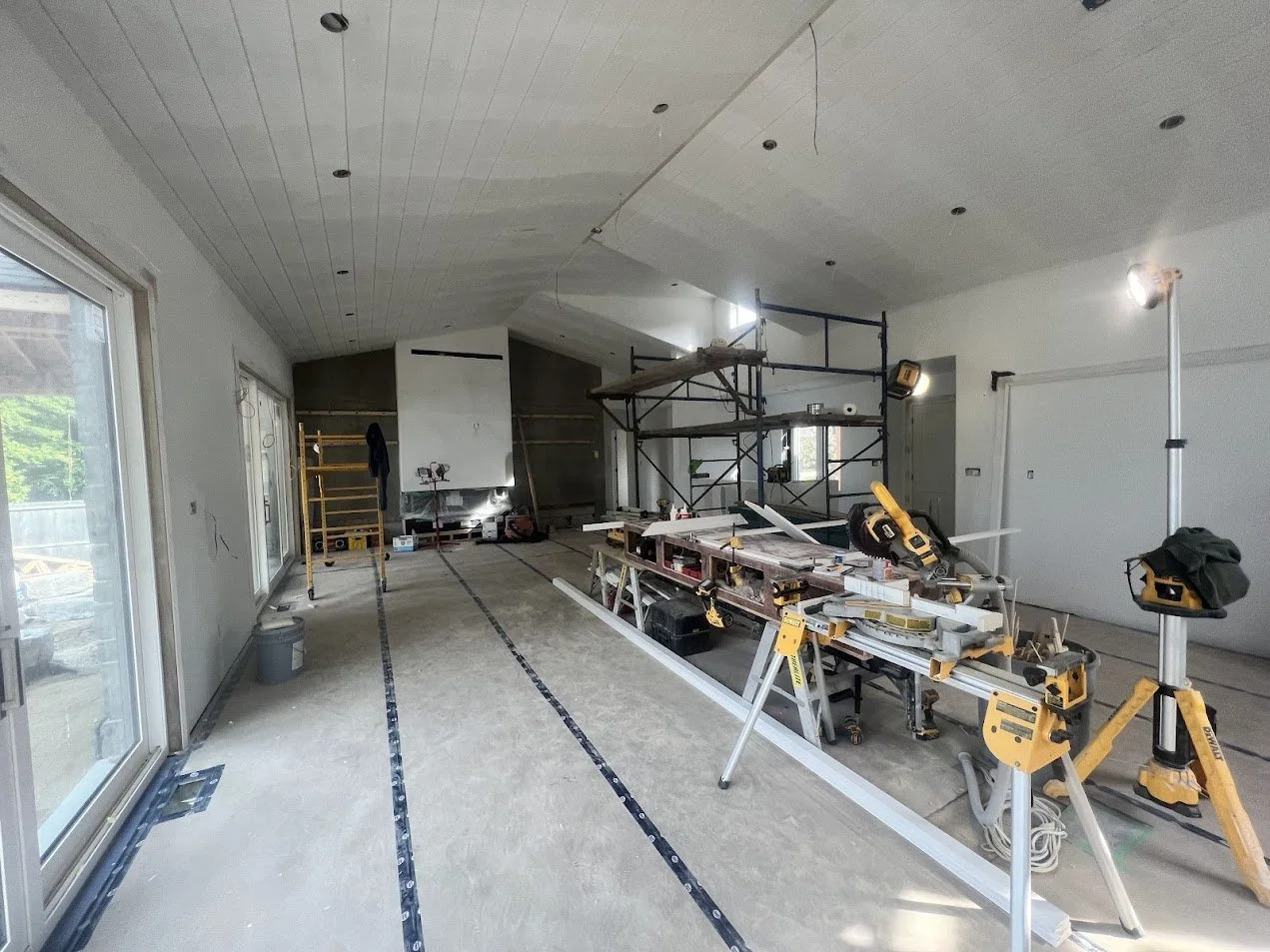 Interior of a house under construction with scaffolding, construction tools, and partially installed fixtures, including large windows and white walls.