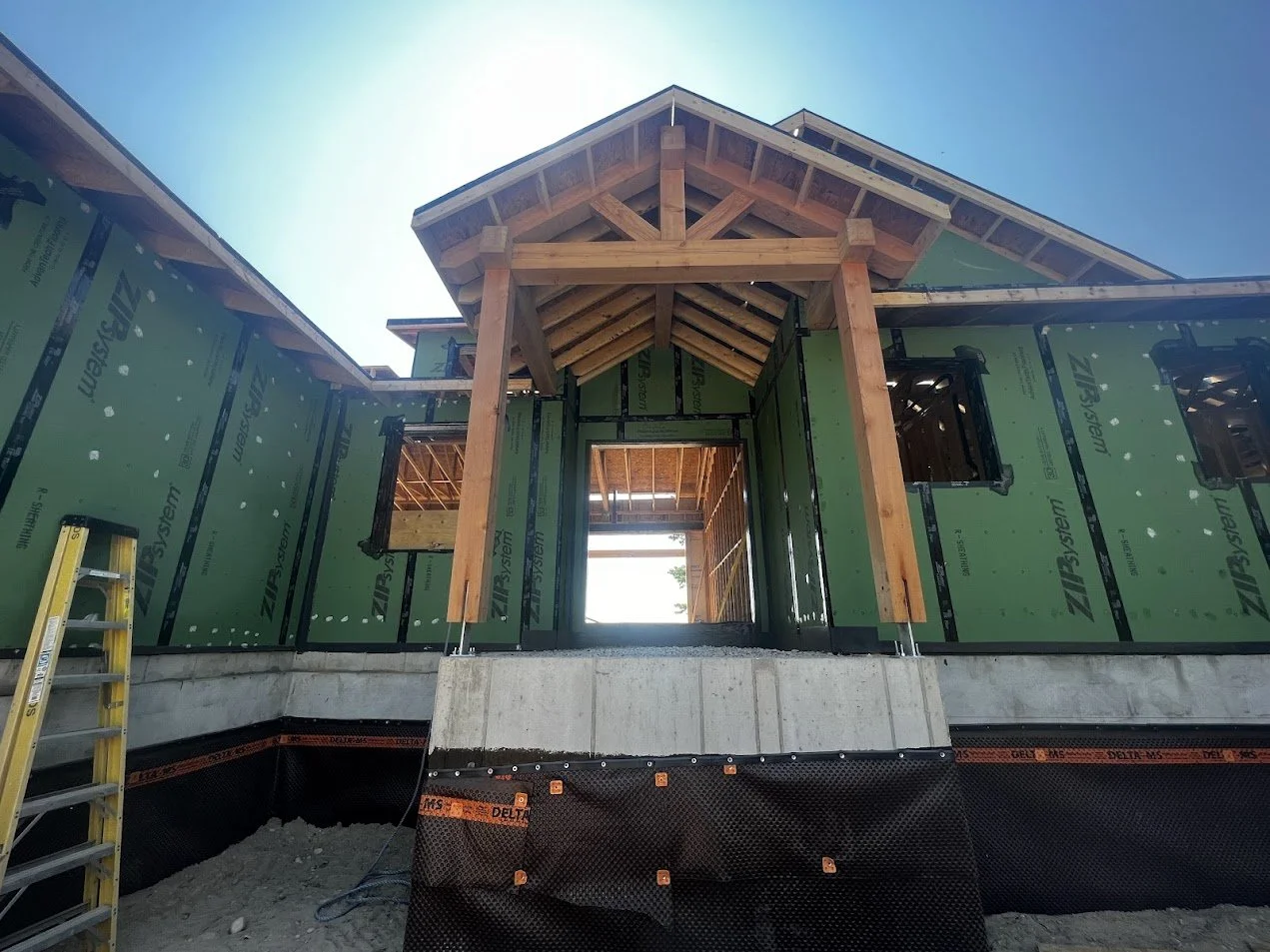 Construction site of a house with green sheathing and exposed wooden framing, stairs leading up to an unfinished entrance, and a ladder on the left side, under clear blue sky.
