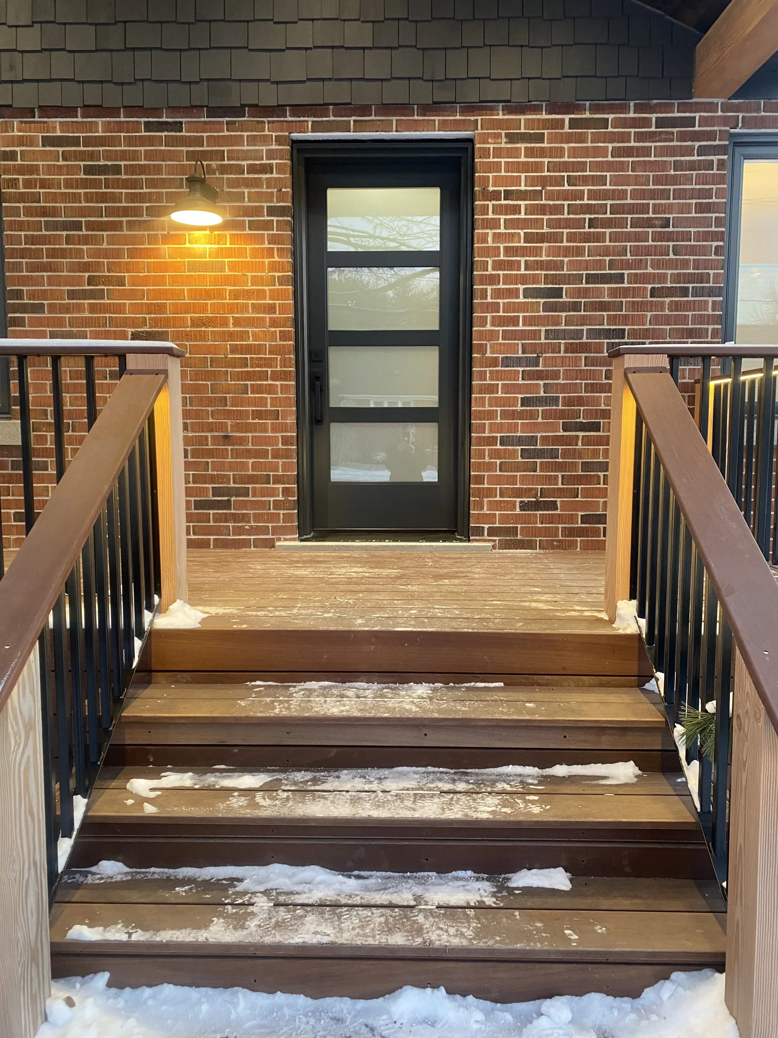 Front porch with wooden stairs, black metal railings, brick wall, a black-framed glass door, warm yellow wall light, snow on the stairs, and a tree reflection in the window.