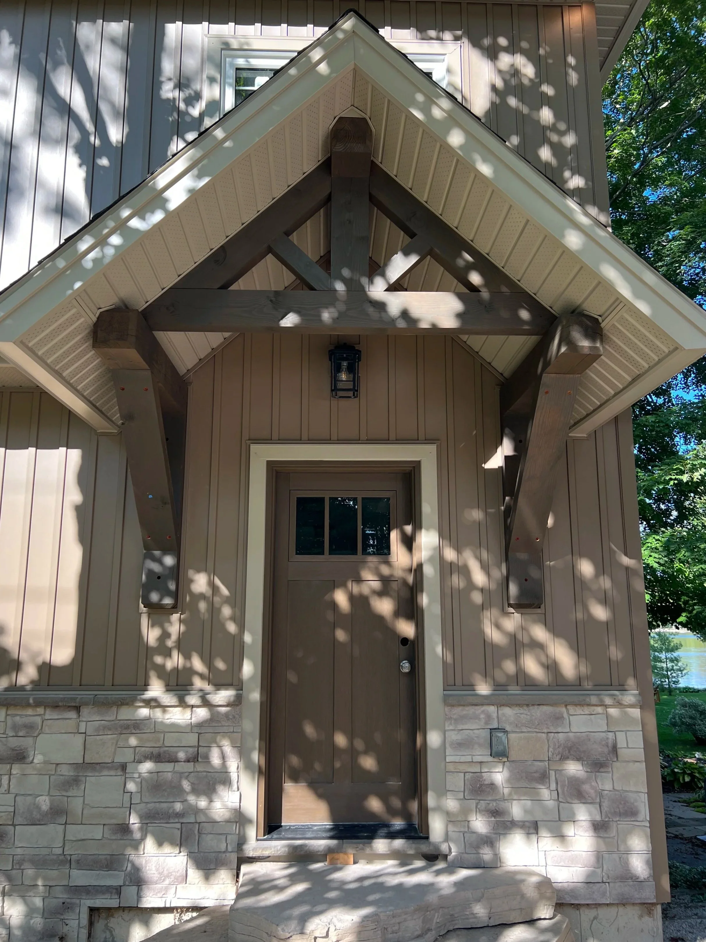 Front entrance of a house with a small porch, wooden door, and stone foundation. The porch is covered by a gabled roof with decorative support beams, and there is a hanging light fixture above the door. Shadows of tree branches are cast on the wall and roof.
