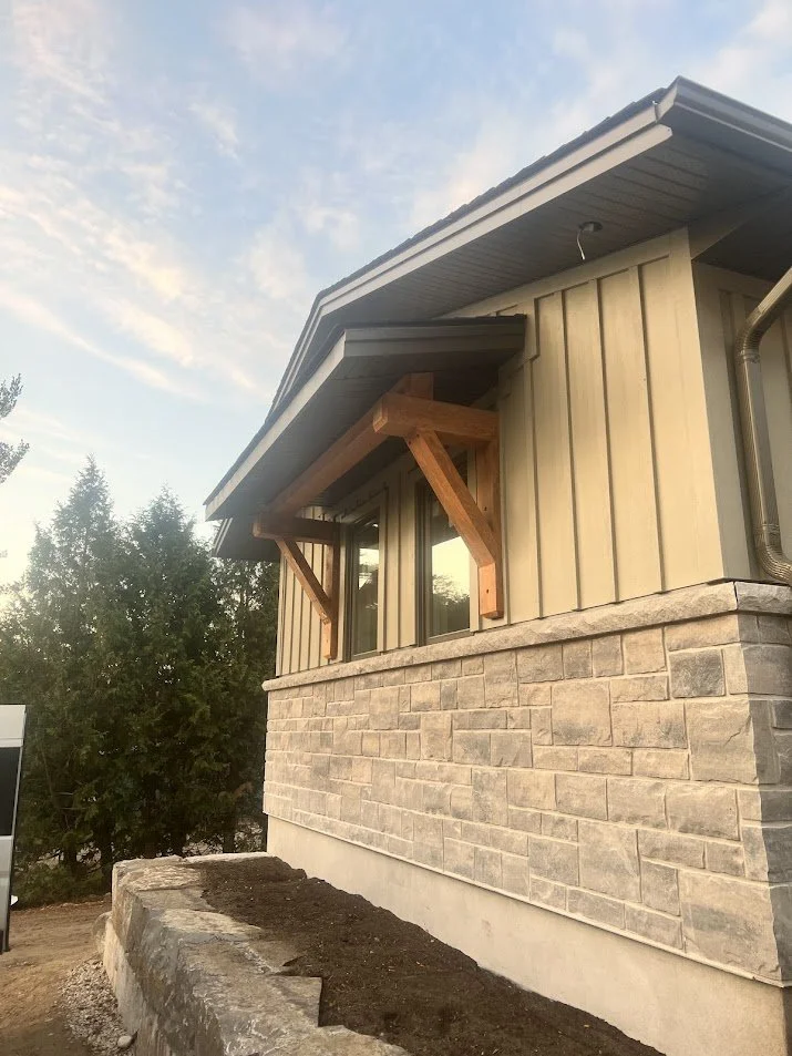 Close-up of a house with vertical beige siding, stone veneer foundation, and a small deck with wooden support brackets under the roof overhang, with trees and a partly cloudy sky in the background.