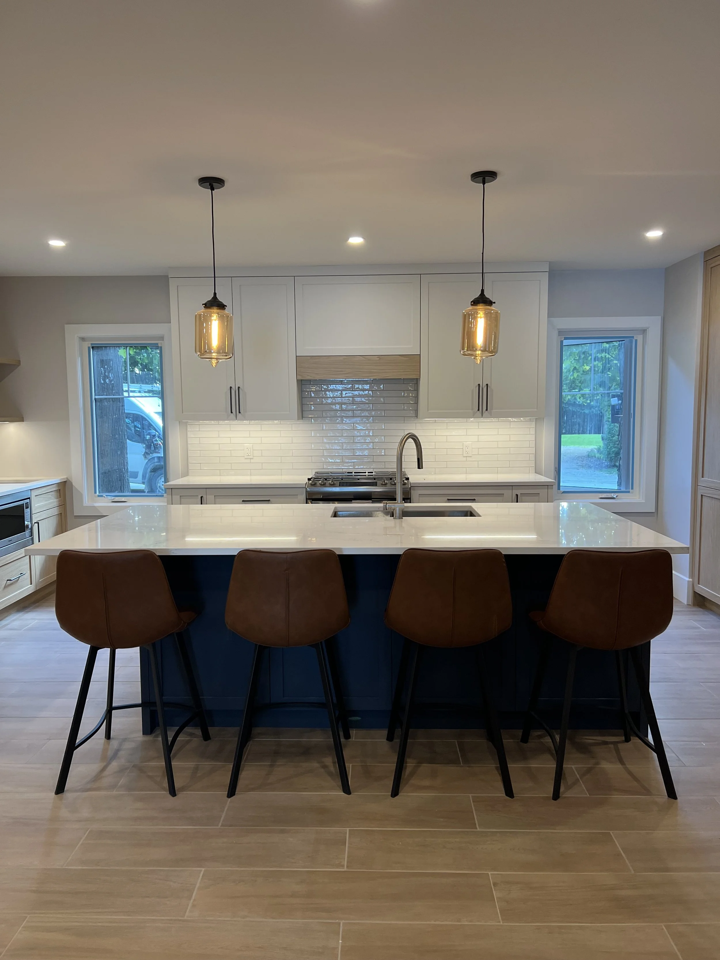 Modern kitchen with white cabinets, a white countertop island with a sink, and four brown barstools. Two pendant lights hang above the island, with windows on either side.