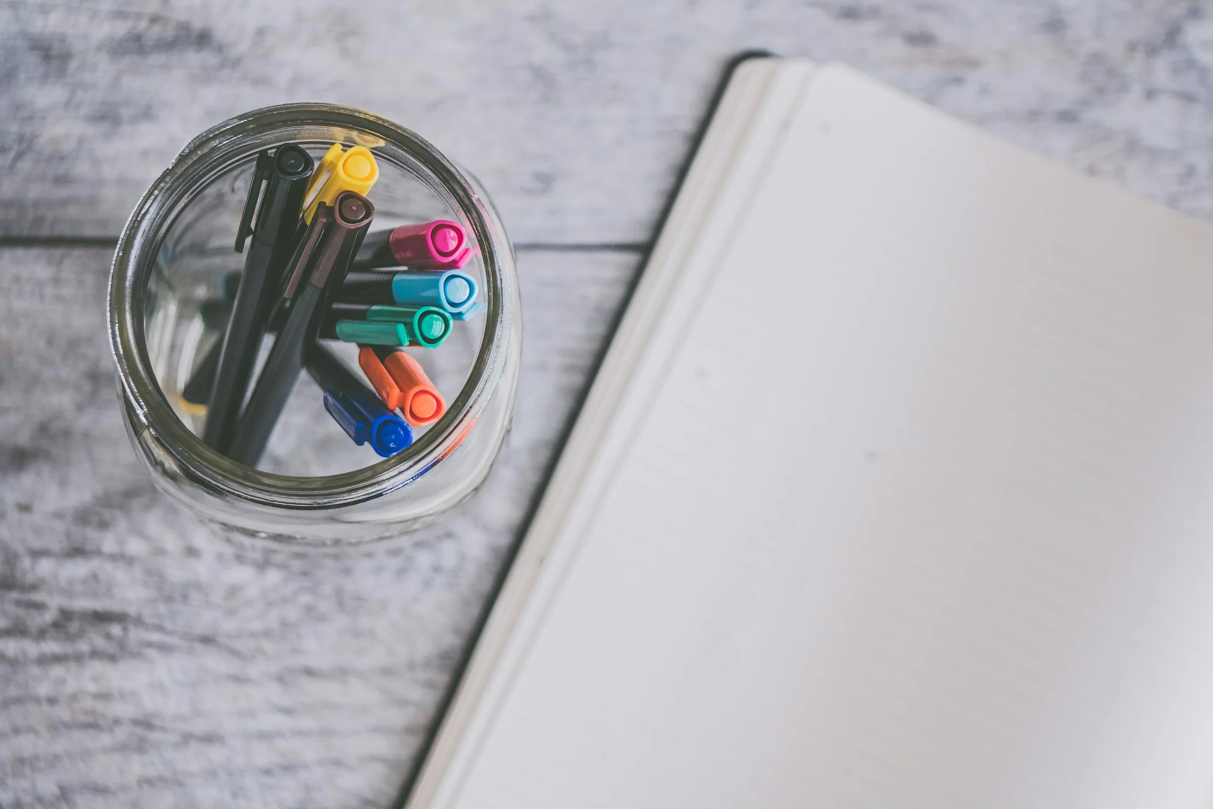 A glass jar filled with colorful markers placed next to a blank notebook on a wooden surface.