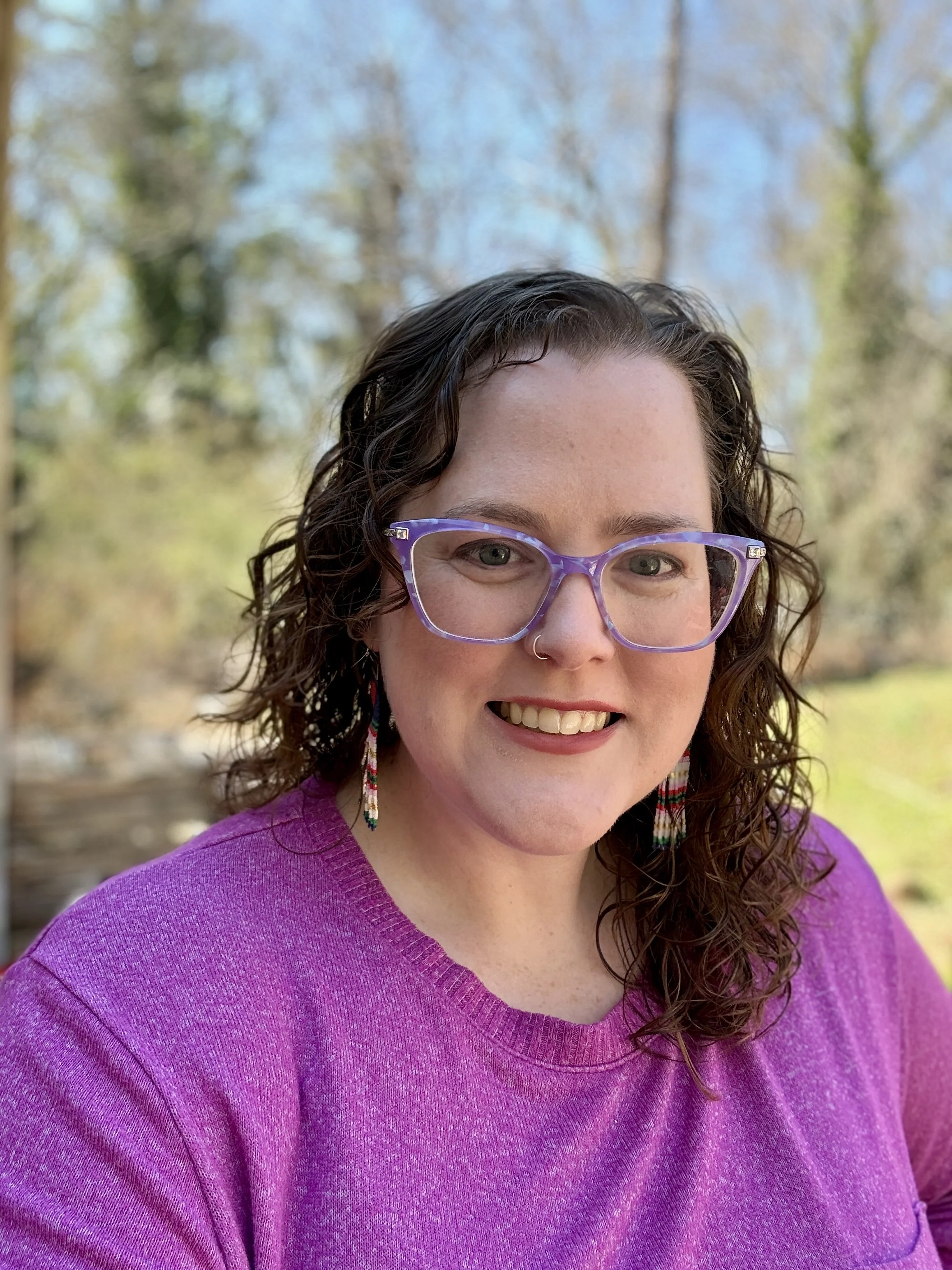 A woman with curly brown hair, purple glasses, and a nose piercing wearing a purple sweater, smiling outdoors with trees in the background.