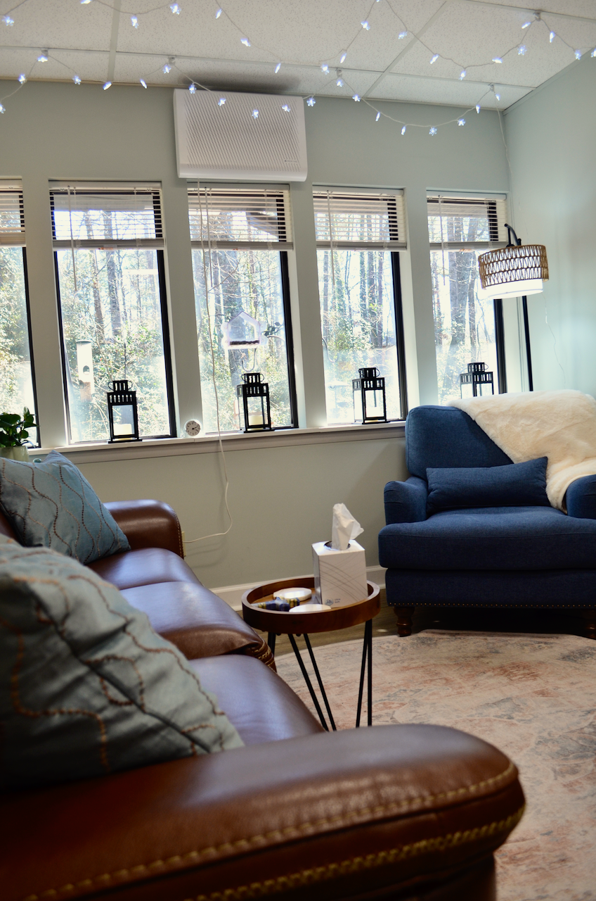 Cozy office space with large windows, blue and brown sofas, decorative pillows, a small round side table with tissue box, string lights on the ceiling, and a view of trees outside.