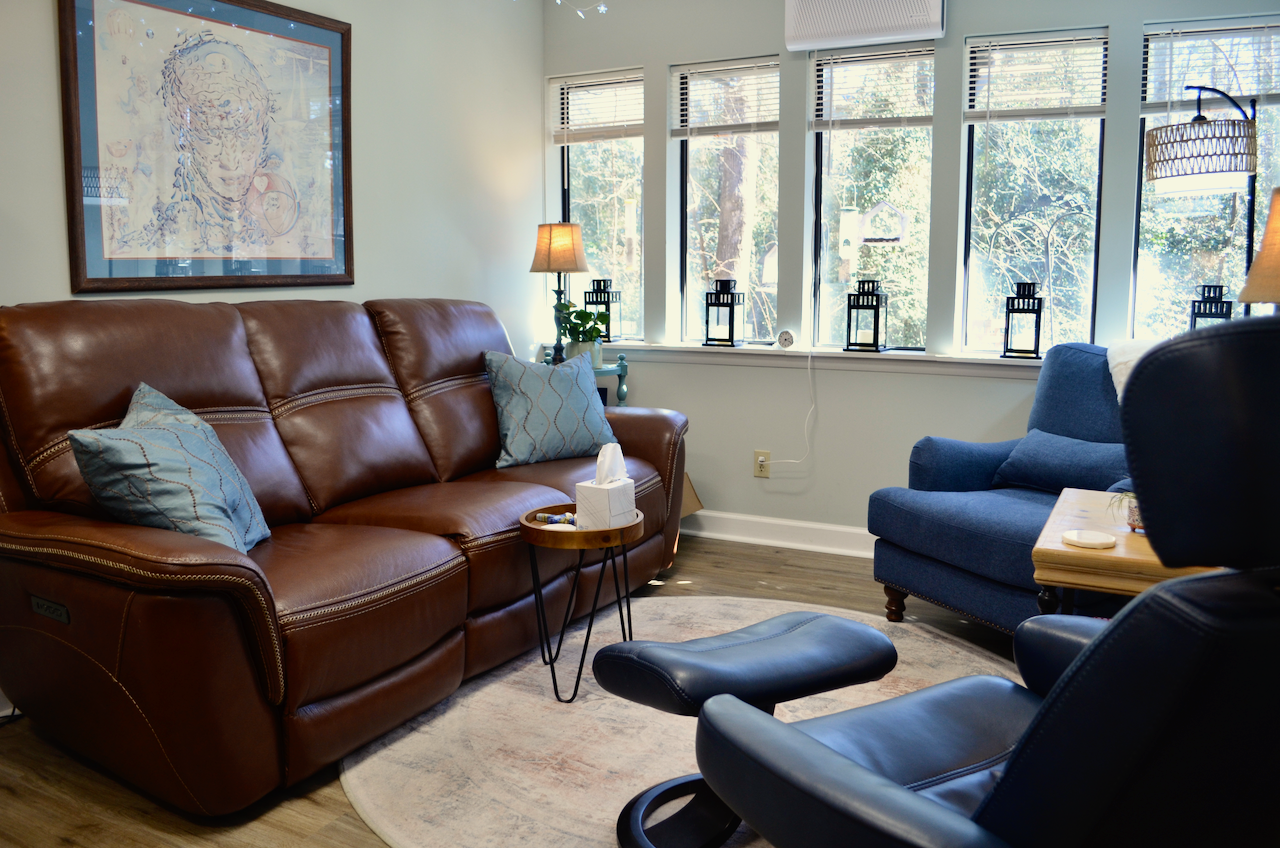 Cozy office space with a brown leather sofa, blue armchair, side table with tissue box, window sill with lanterns, and a large framed artwork on the wall.