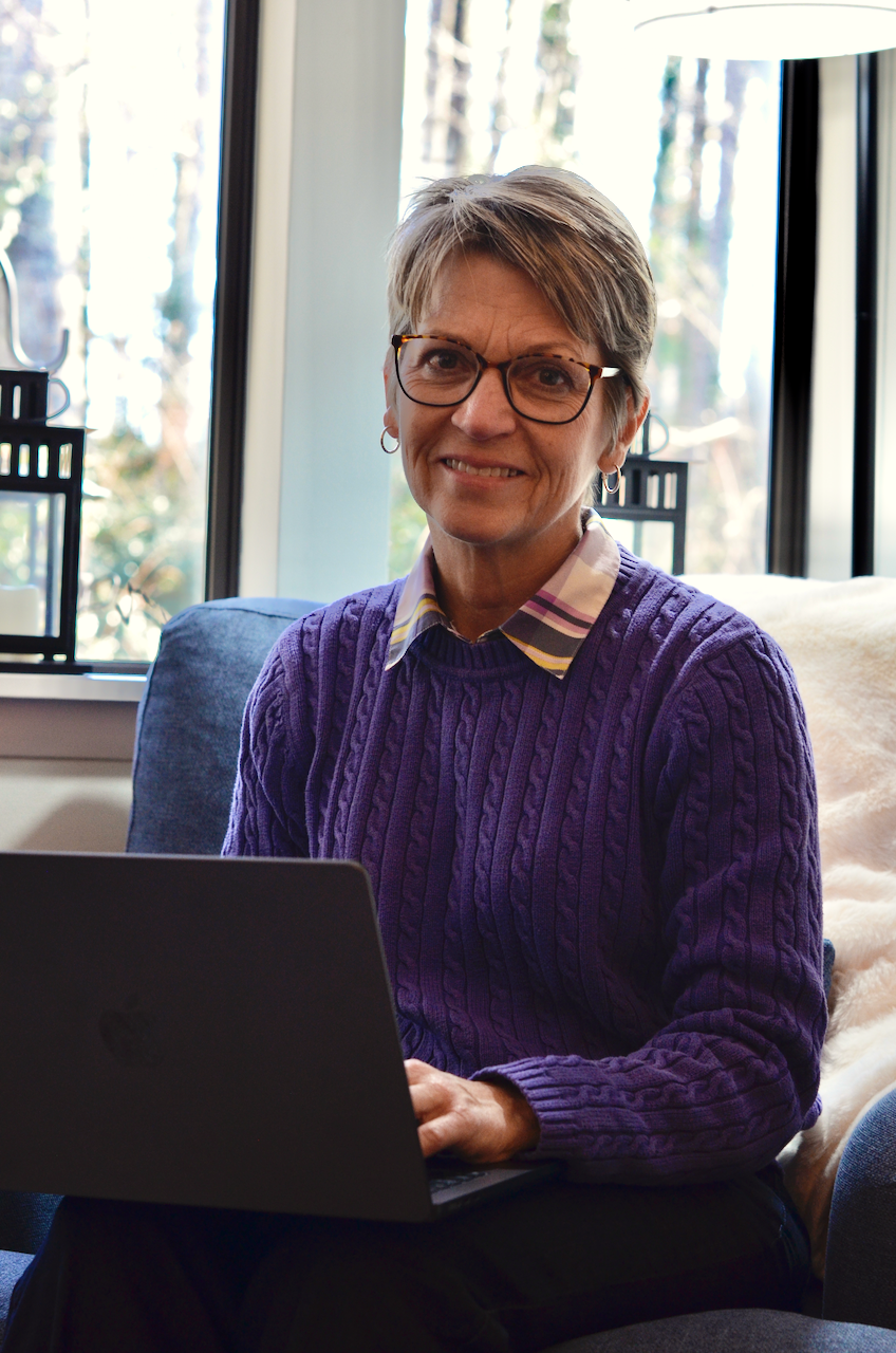 A smiling woman with short gray hair and glasses working on a laptop indoors, with a window showing trees outside.