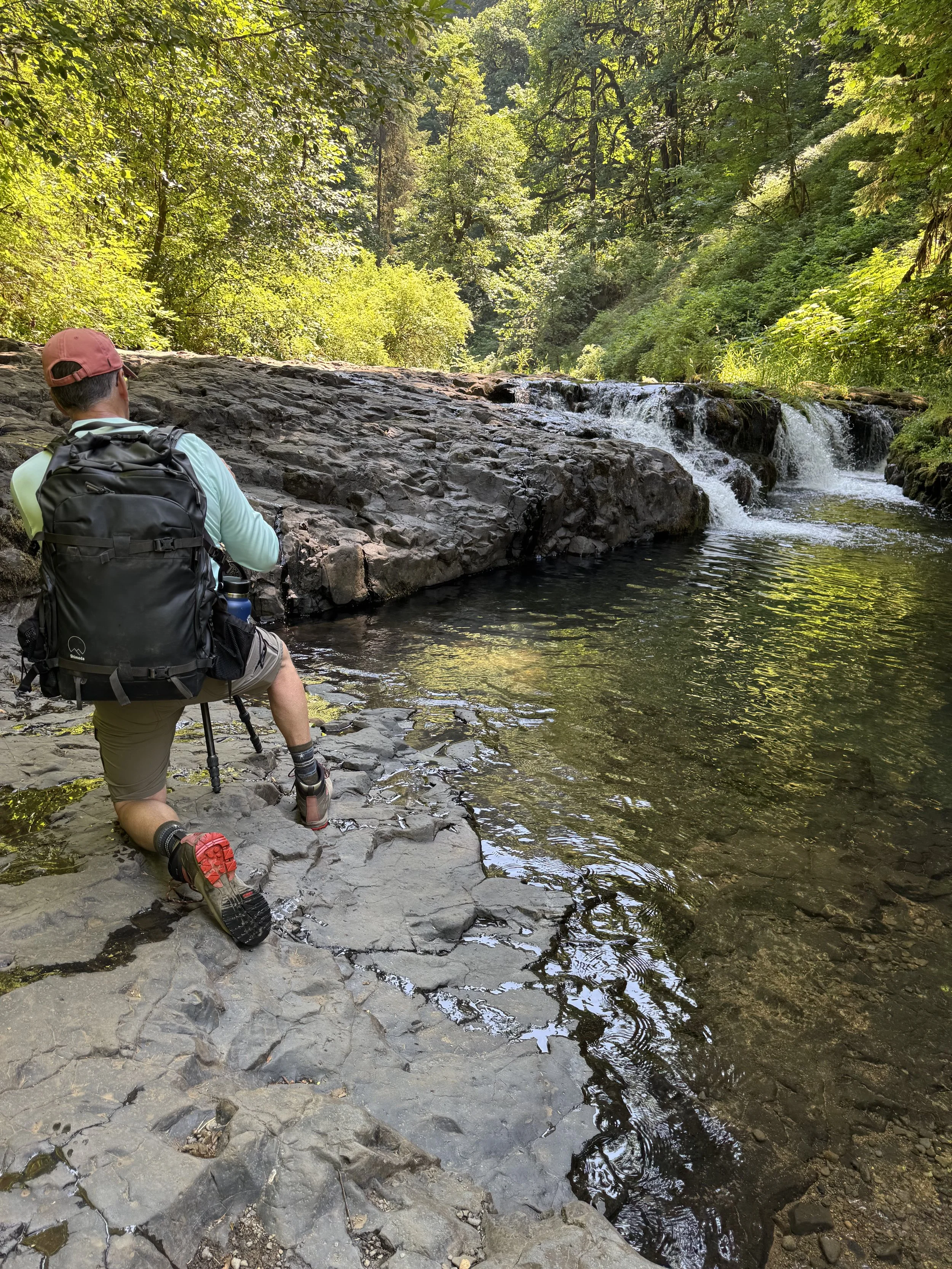 A hiker kneeling on rocks beside a river with a small waterfall, surrounded by lush green trees in a forest setting.