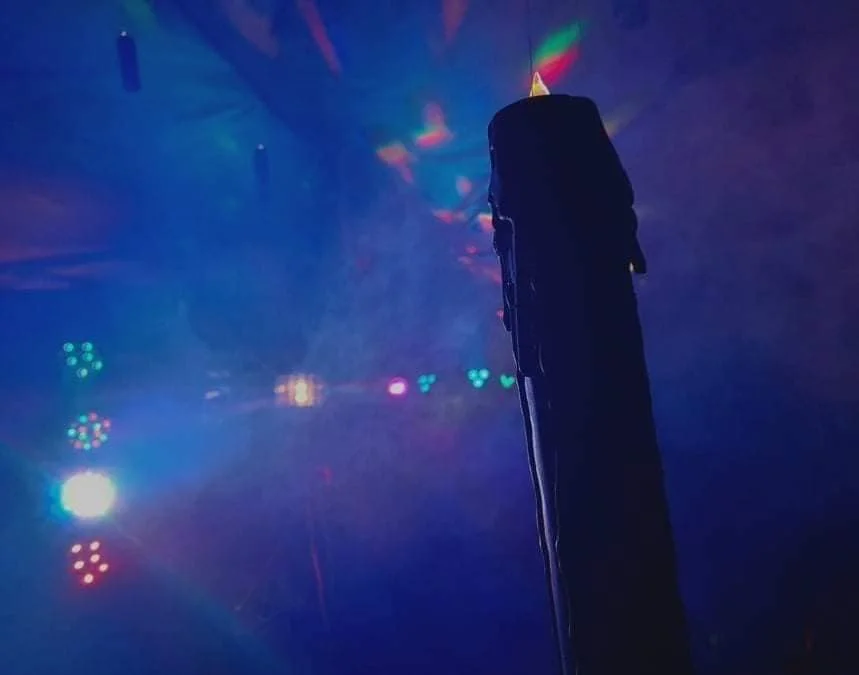 A black object, possibly a microphone or similar device, is standing upright in a dark room illuminated by colorful stage lights, creating a vibrant, foggy atmosphere with blue, purple, and green hues.