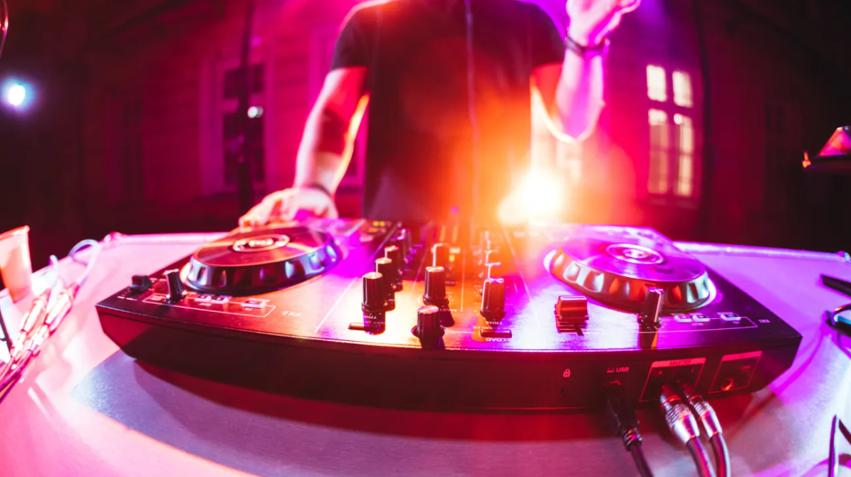 DJ in a dark room with pink and purple lighting, standing behind a DJ controller with various knobs and turntables.