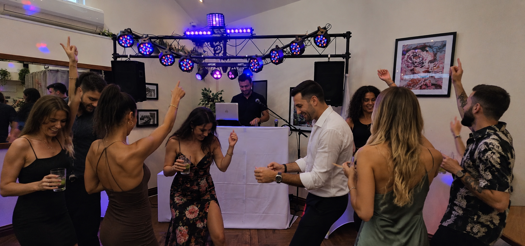 People dancing and enjoying music at a party with a DJ in the background, colorful lighting, and some framed artwork on the wall.