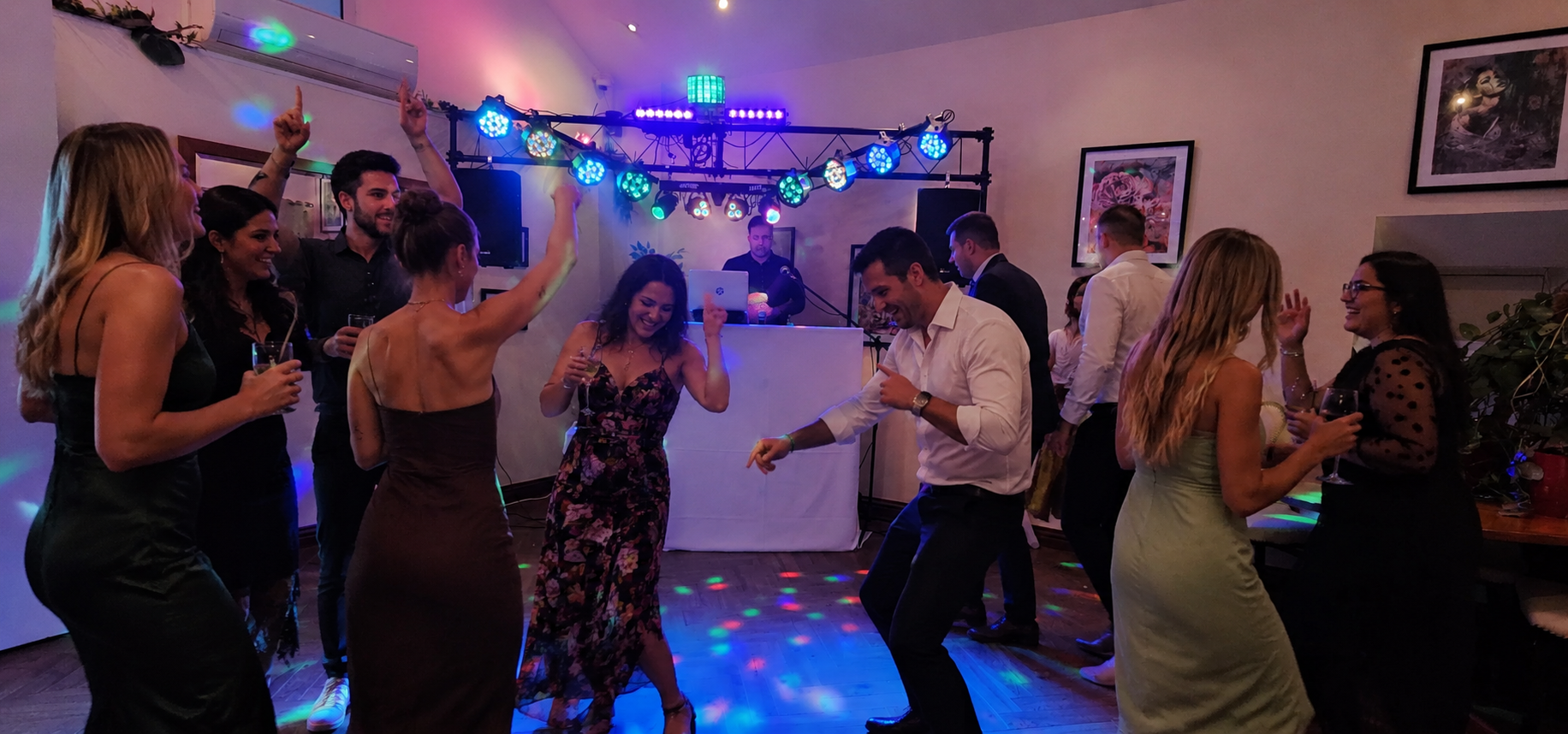 People dancing and socializing at a lively party with a DJ in the background and colorful lighting.