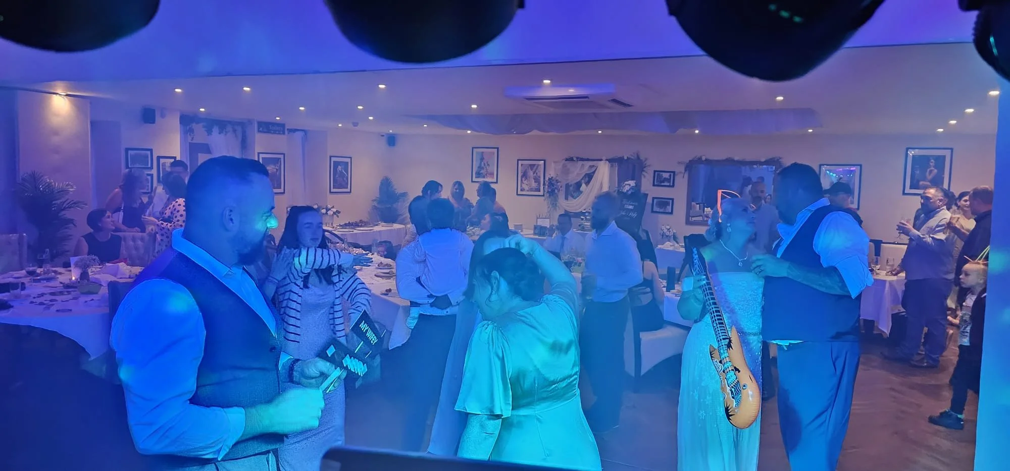 People socializing and dancing at a party or celebration in a decorated room with framed pictures on the walls, tables with food, and a woman holding a guitar visible.