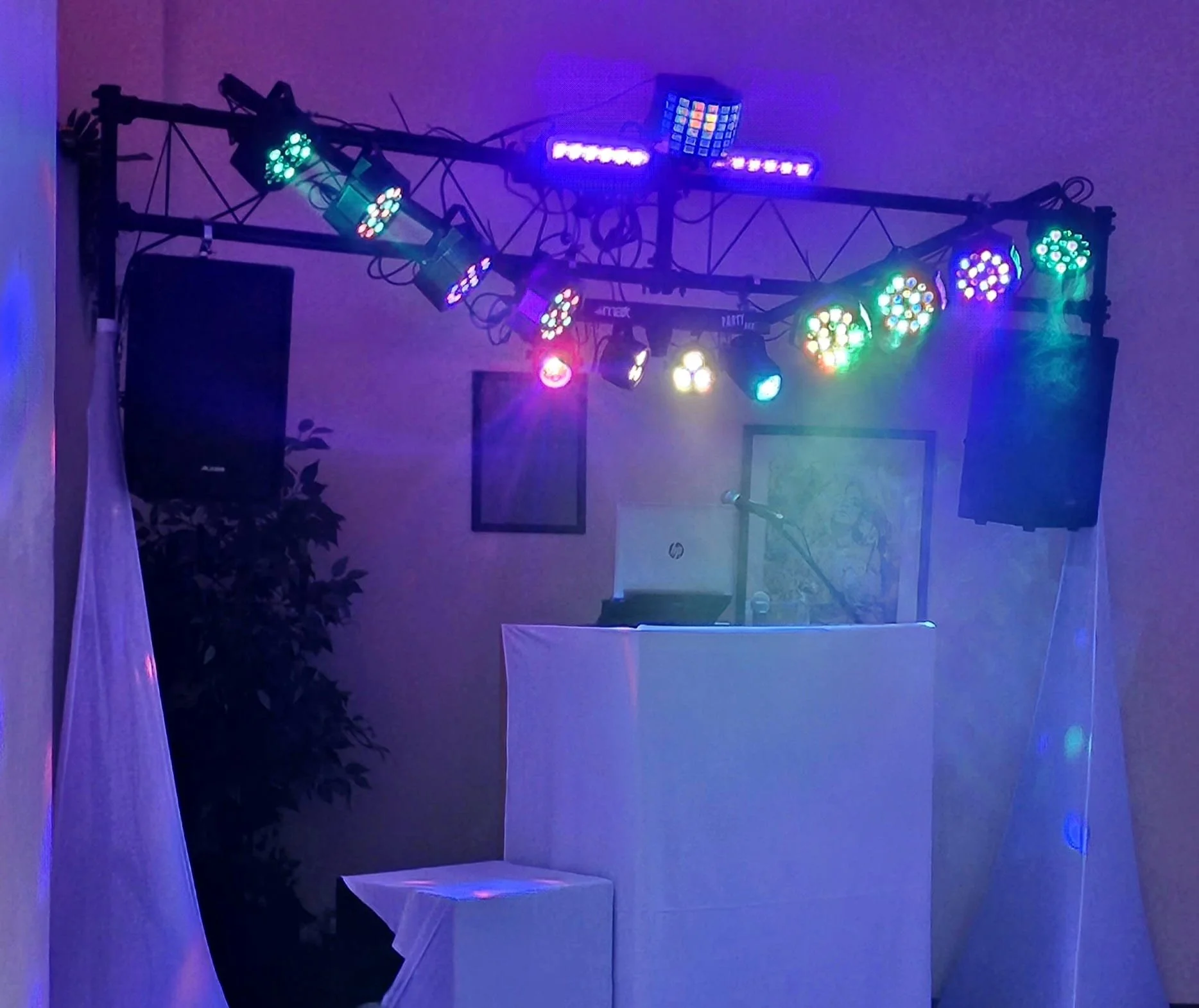 DJ booth with colorful stage lights, speakers, microphone, and white drapery in a dimly lit room