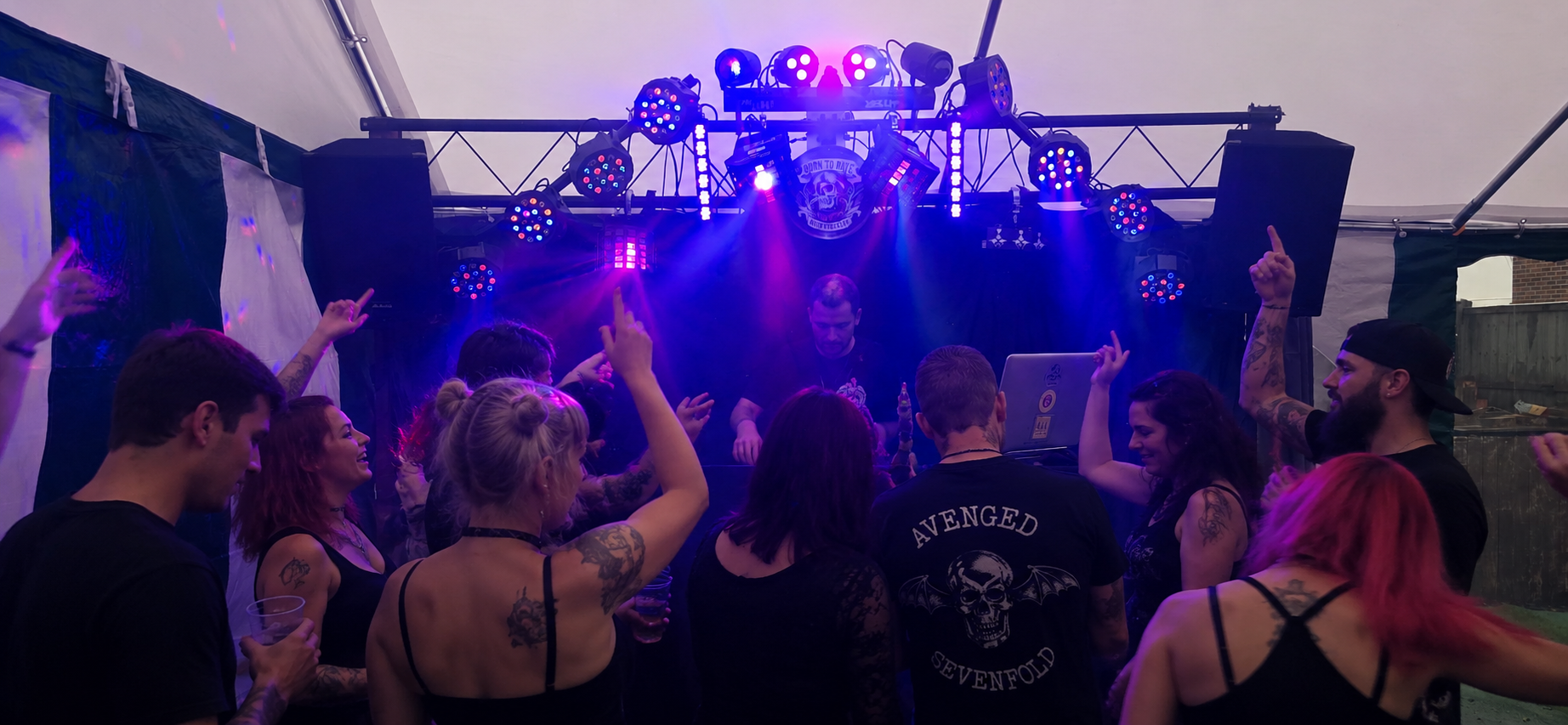 People dancing and enjoying music at a live DJ event in a tent with purple and pink lighting.