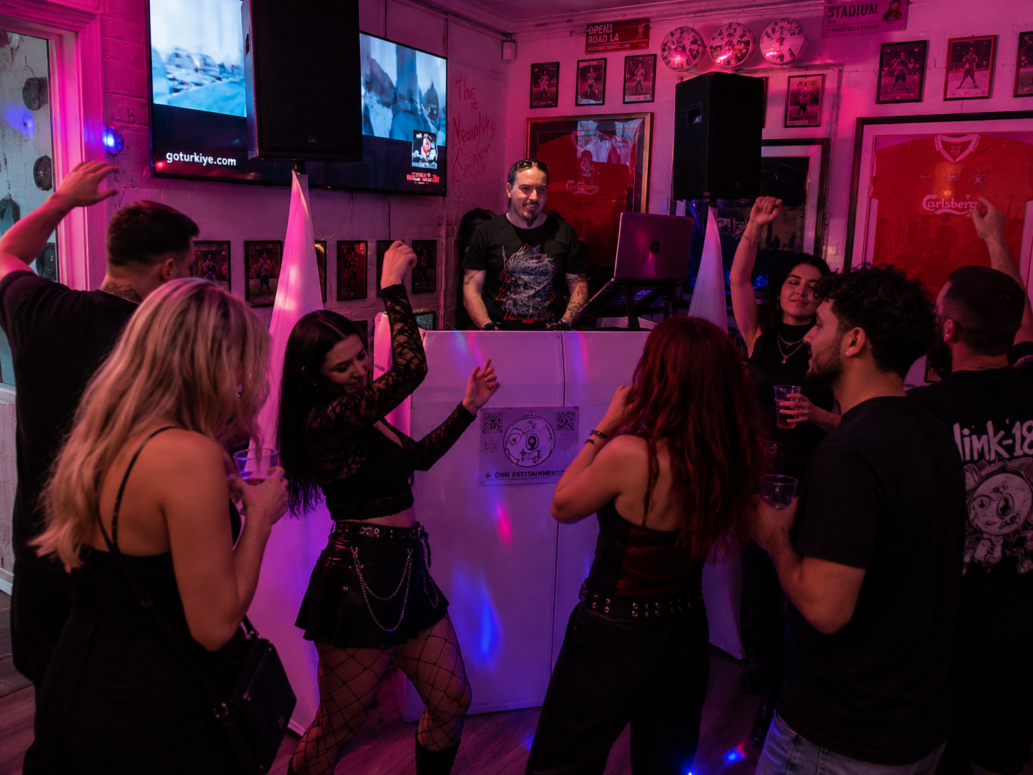 People dancing and socializing inside a nightclub with a DJ playing music. The room features pink and purple lighting, framed sports jerseys, and posters on the walls. Some individuals are raising their hands, holding drinks, and enjoying the music.