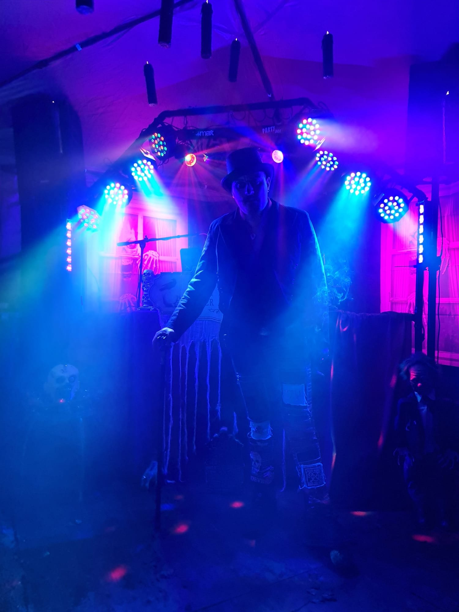 Person in dark clothing and a top hat standing under colorful stage lights with purple, blue, and pink hues, in a decorated setting with Halloween-themed decorations such as skulls and skeletons.