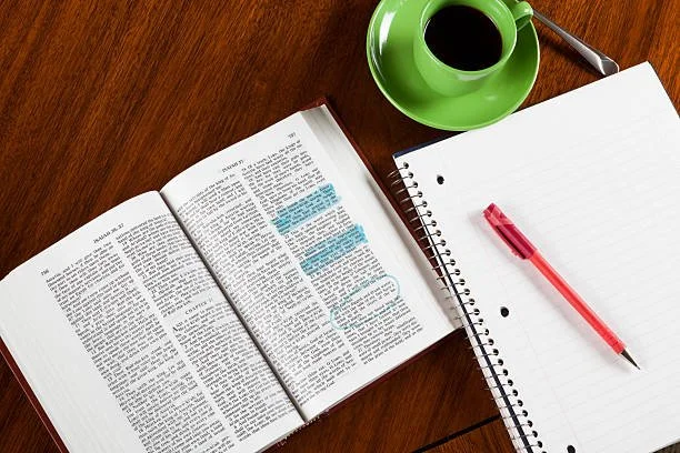 Open book with highlighted text, blank spiral notebook with a red pen, and a green cup of coffee on a wooden table.