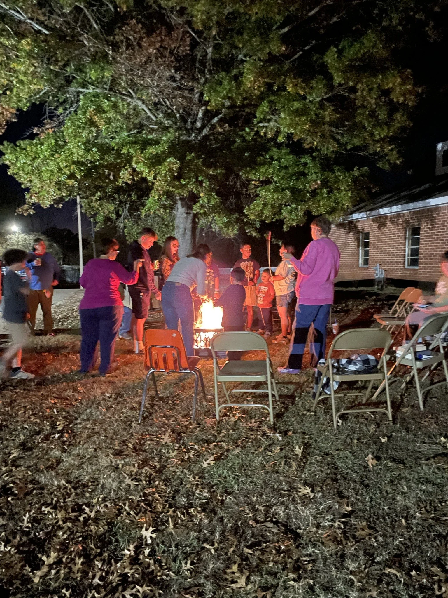 Group of children and adults gathered around a small campfire at night in a backyard yard near a large tree and brick building, some sitting on folding chairs.