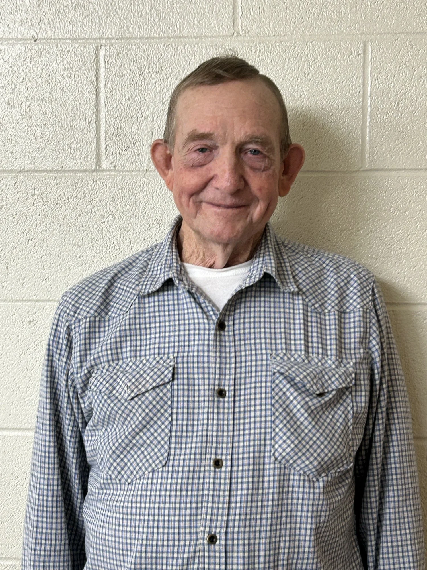 A young man sits on a beige bench against a beige wall, wearing a gray polo shirt and cream-colored pants, looking to the side with a contemplative expression.