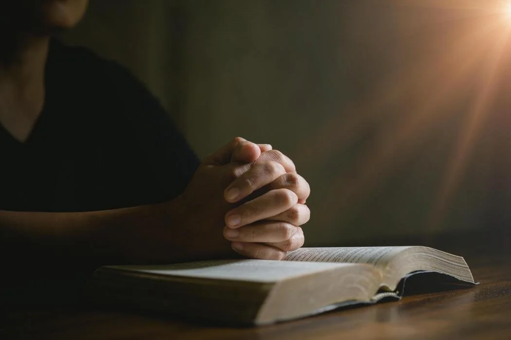 Person praying with hands clasped over an open Bible, in a dimly lit room.