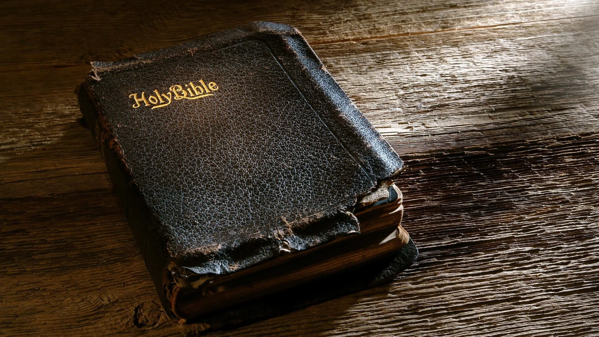 A worn, black leather-bound Bible with gold lettering on the cover resting on a textured wooden surface.