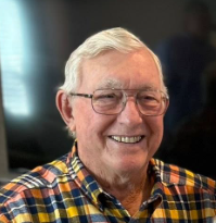 An elderly man with white hair and glasses smiling, wearing a colorful plaid shirt.