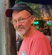 A man with glasses and a beard wearing a red cap and red t-shirt, standing outdoors near a tree in what appears to be a lively area with people and colorful lights.