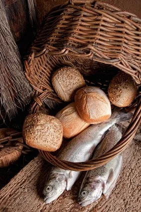 A wicker basket containing five fish and several round bread rolls, placed on a wooden surface.