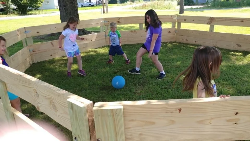 Five children playing with a blue ball inside a wooden fenced area in a park.