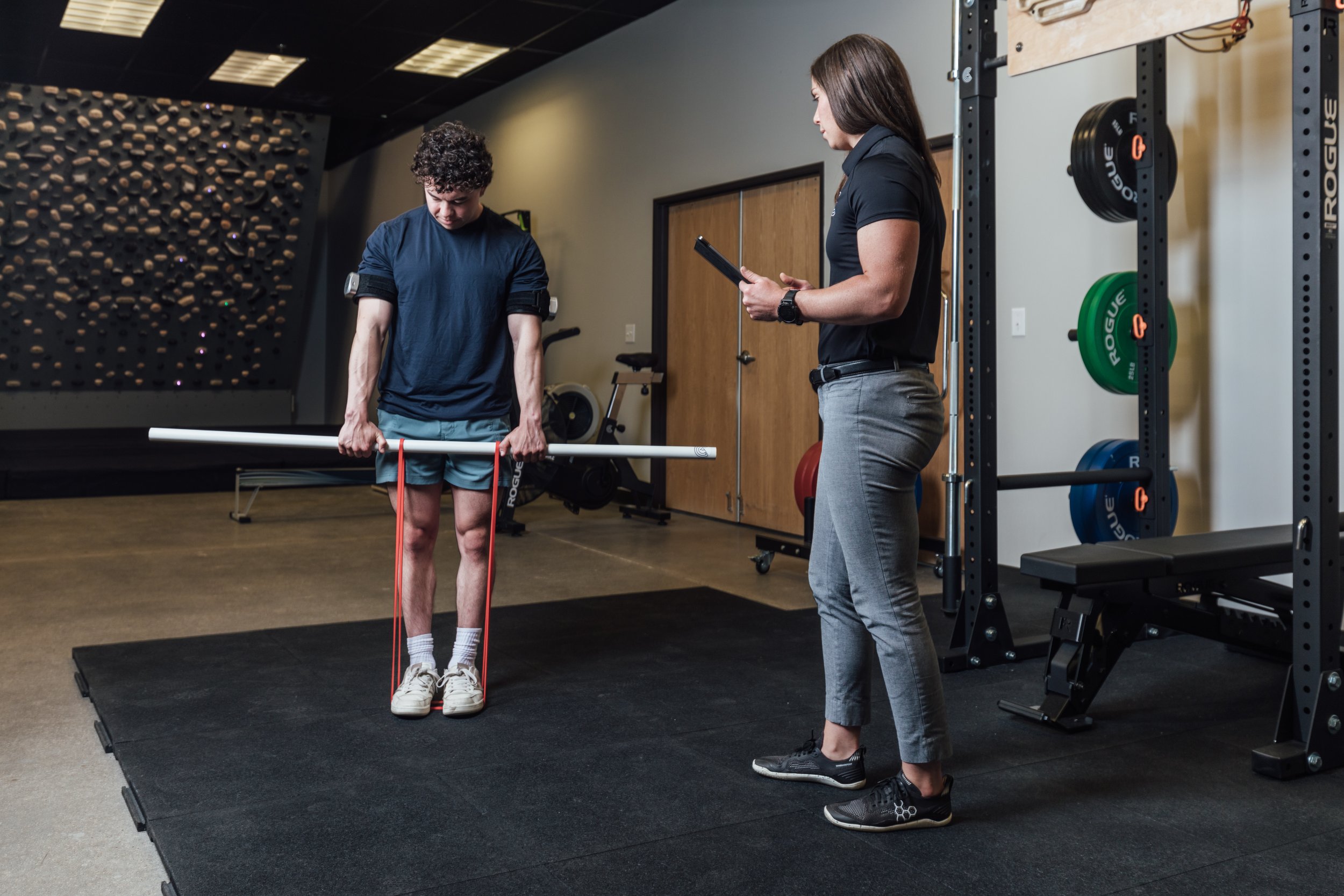 A young man doing a resistance band exercise on his legs while standing on a black mat, with a woman trainer holding a clipboard and observing in a gym with weightlifting equipment in the background.