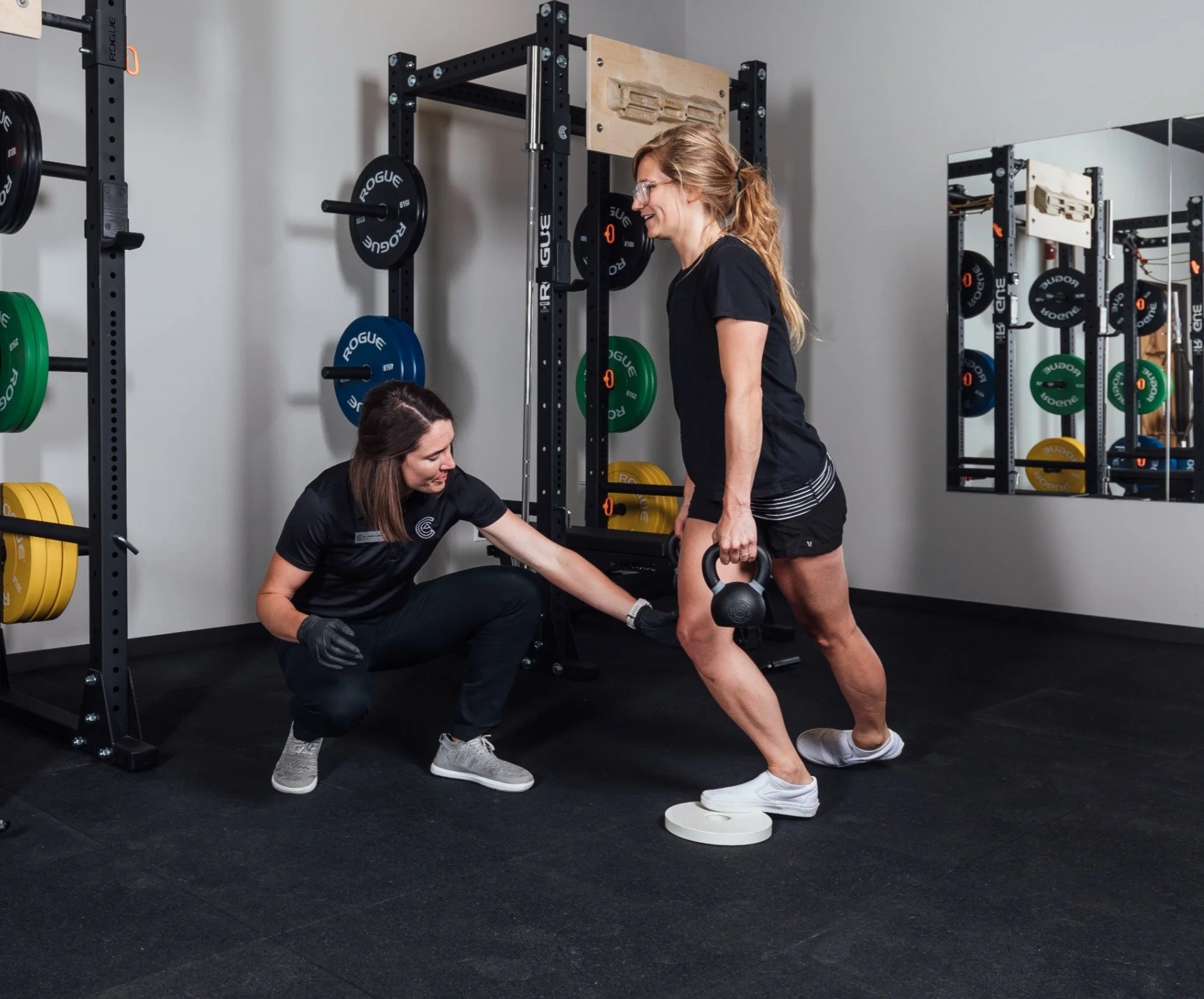 A woman holding a kettlebell doing a squat with a trainer assisting her in a gym.