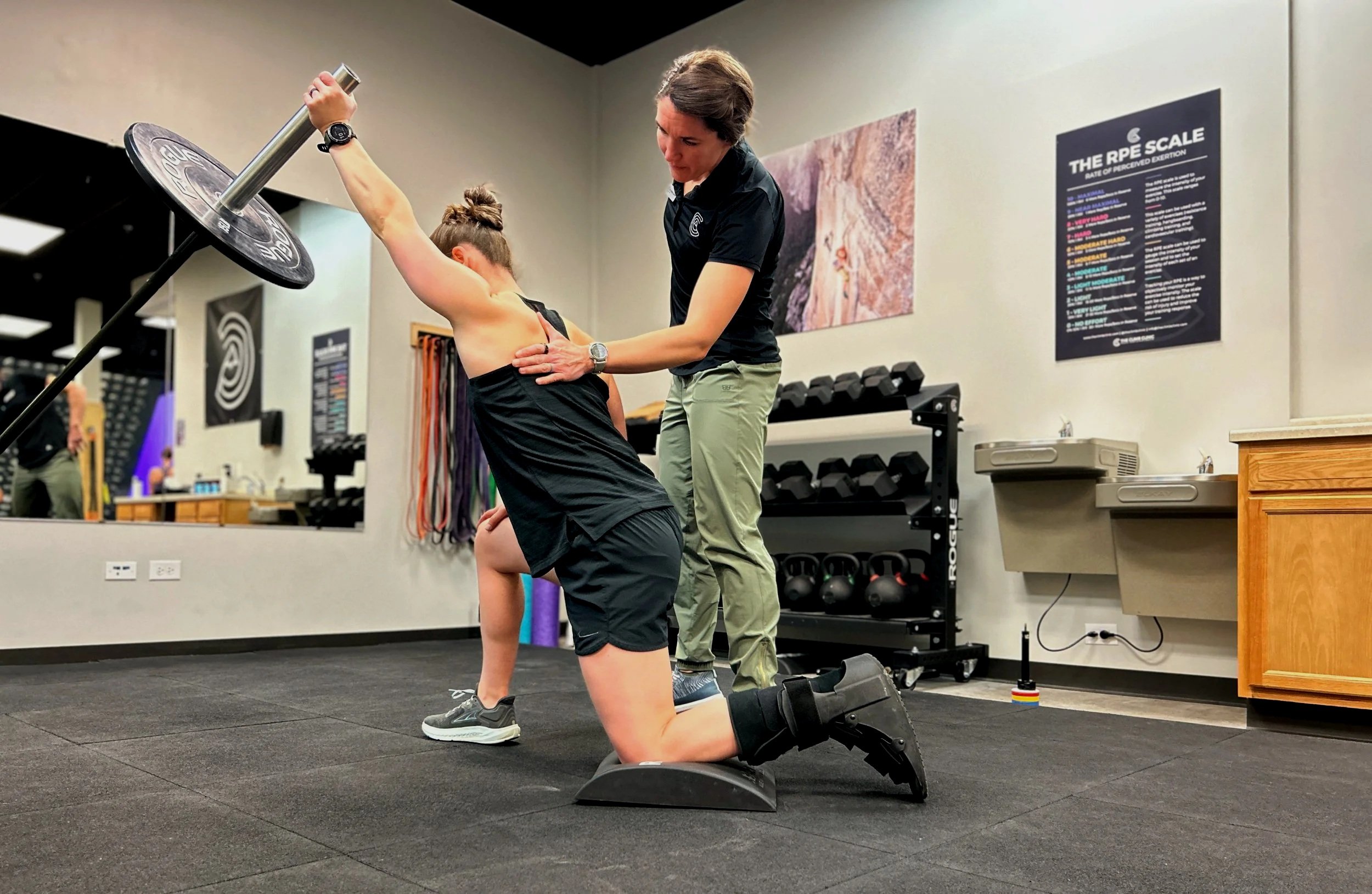 A woman is performing a kneeling exercise with a barbell on her back, assisted by a trainer in a fitness gym.