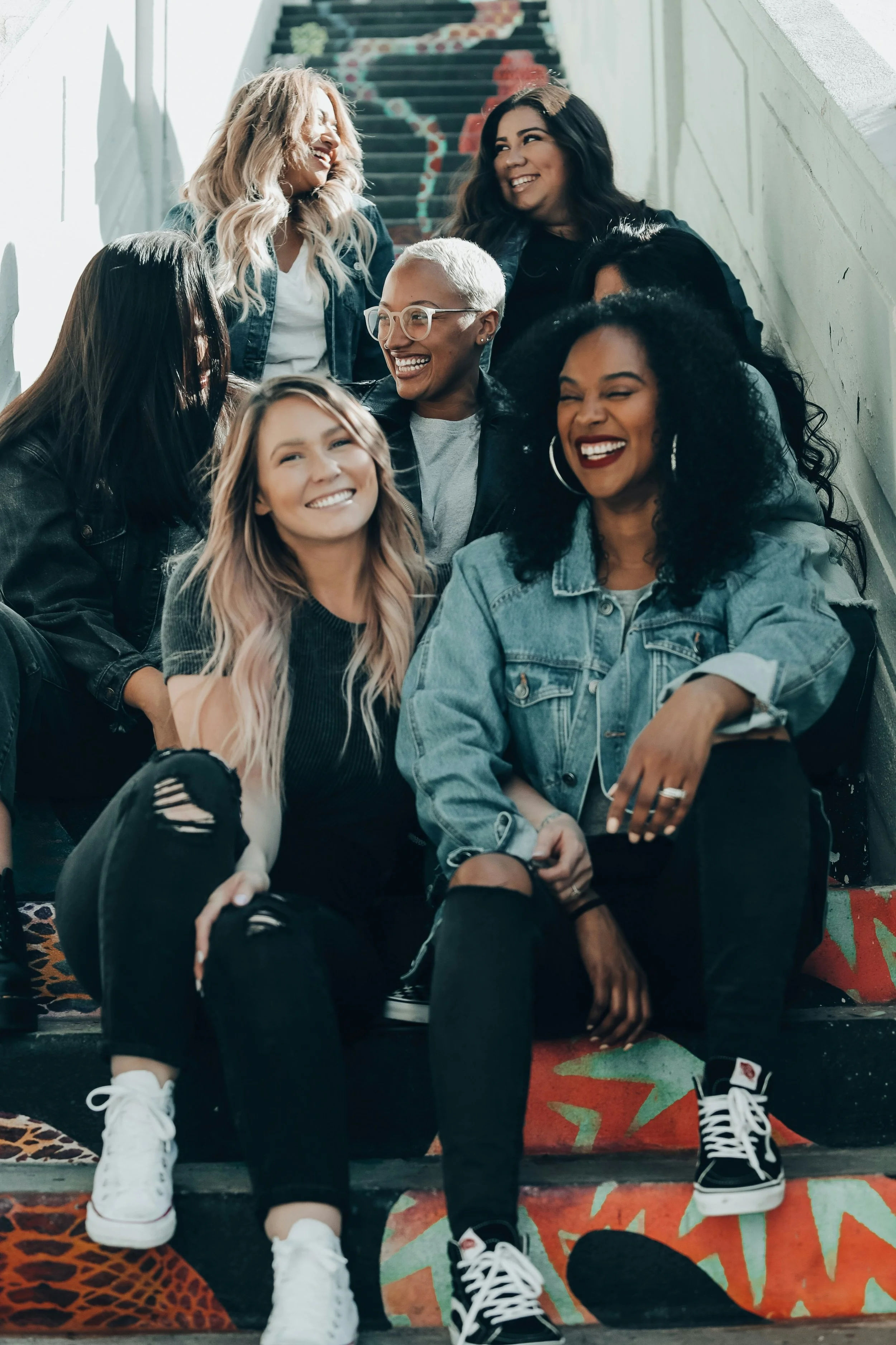 Group of seven women sitting on stairs, smiling and laughing together, with colorful graffiti on the wall and stairs behind them.