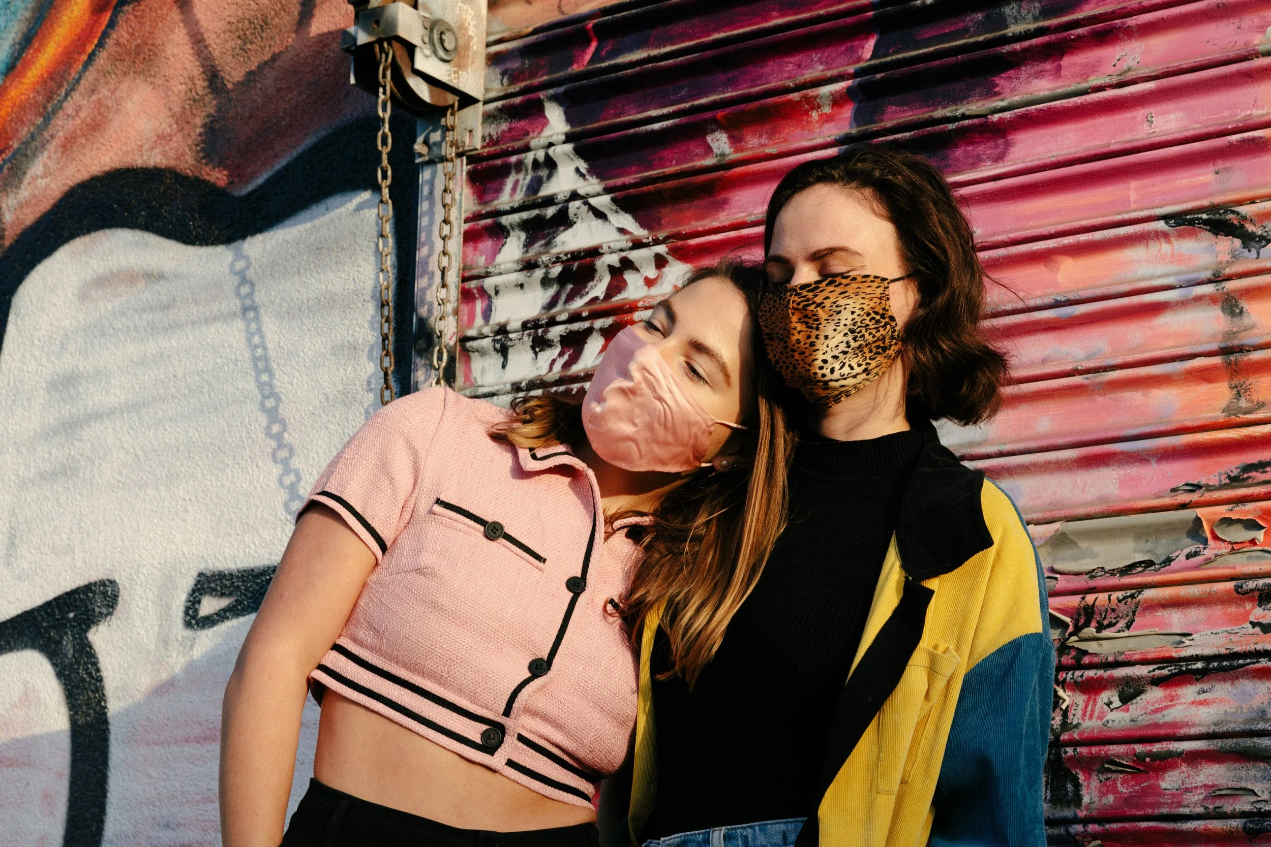 Two young people wearing face masks standing close together against a graffiti-covered metal wall.