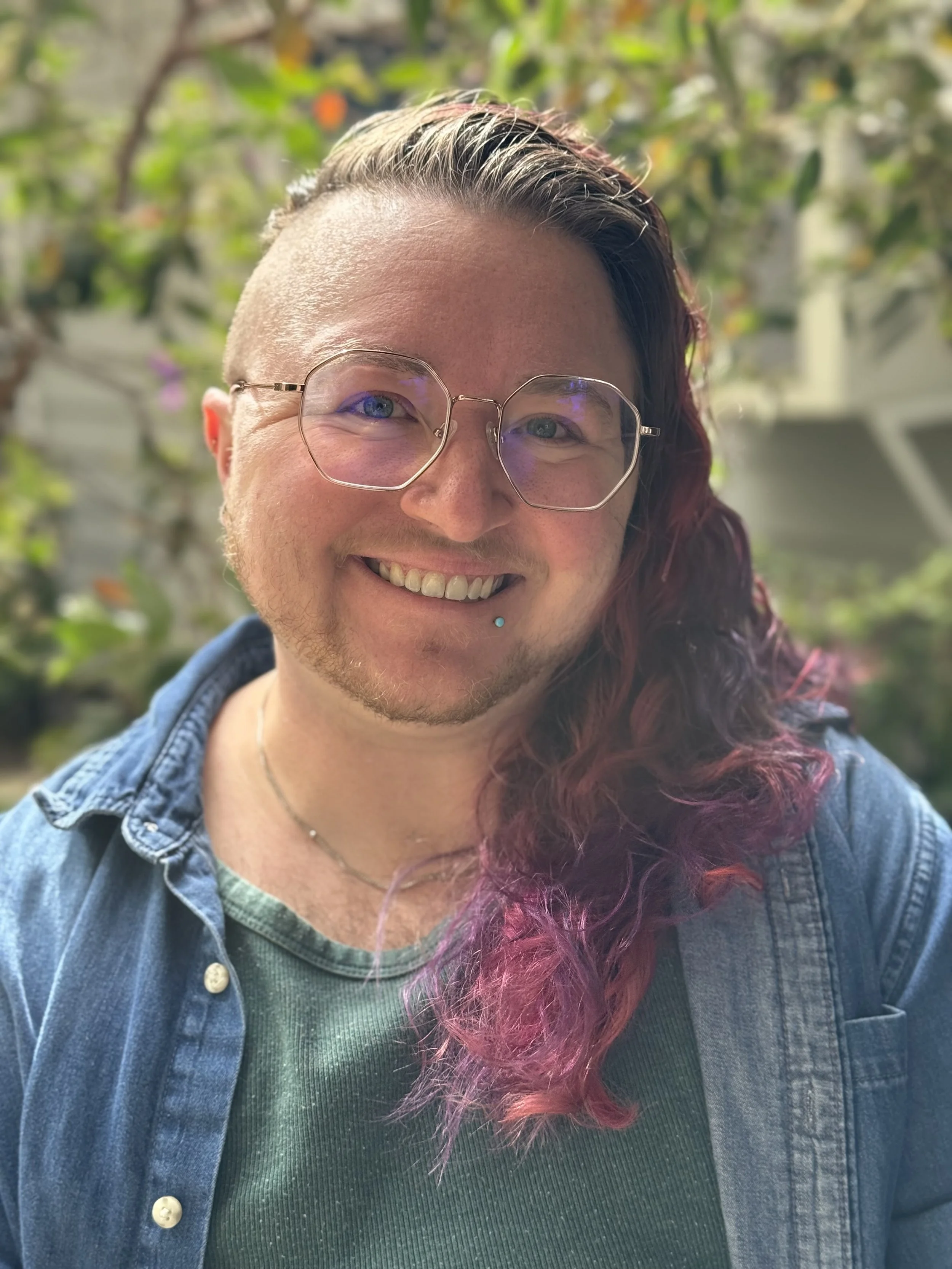 A transgender queer therapist, Jay Klyman, they have glasses and a blue denim jacket, with purple and pink hair, standing outdoors with greenery in the background.