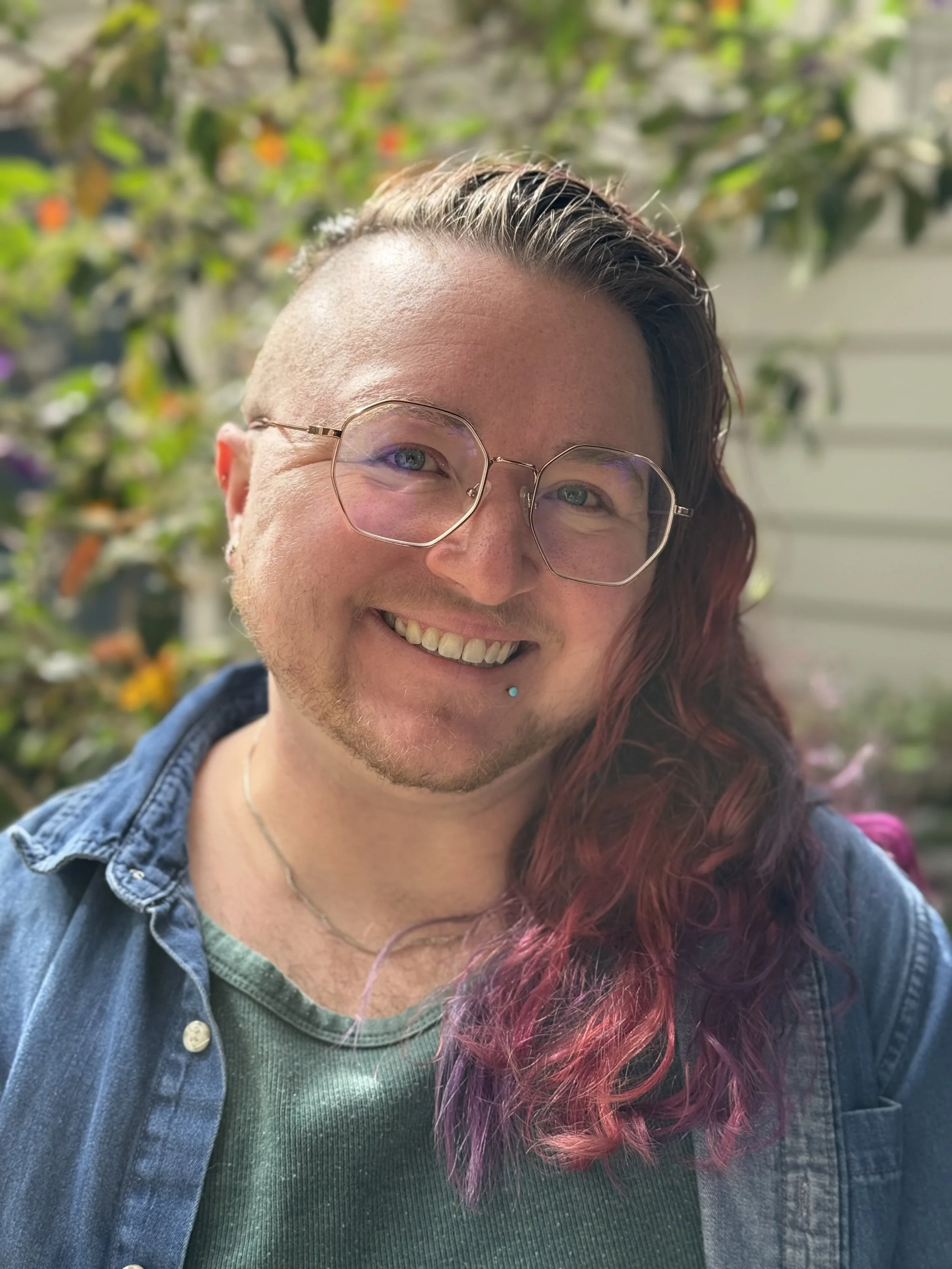 Smiling LGBTQIIA+ person with glasses, a lip piercing, and long hair that is dark at the top and pink at the ends, outdoors with green foliage in the background.