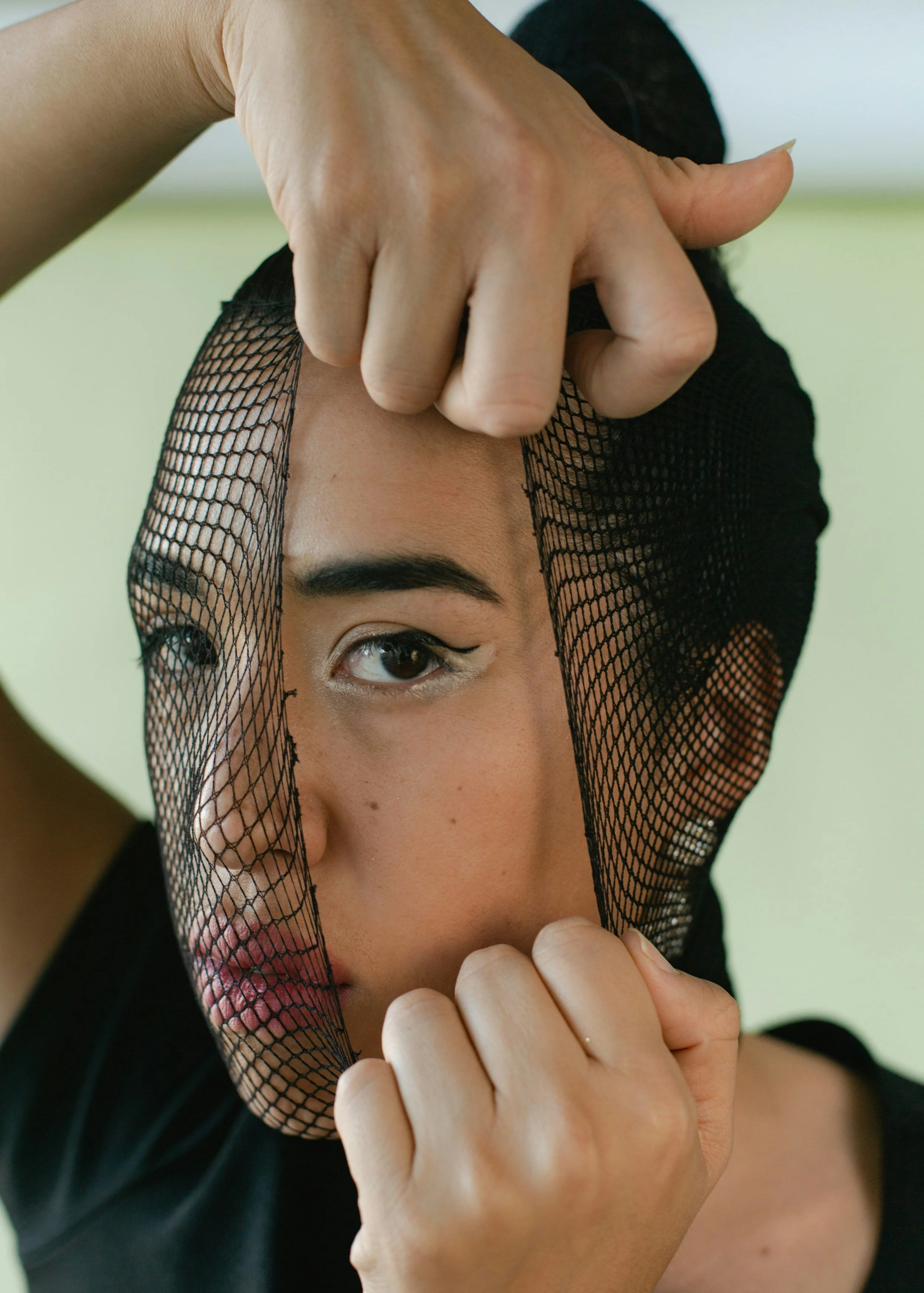 A queer Asian woman peeling back a fish net that is covering her face that has make-up on it
