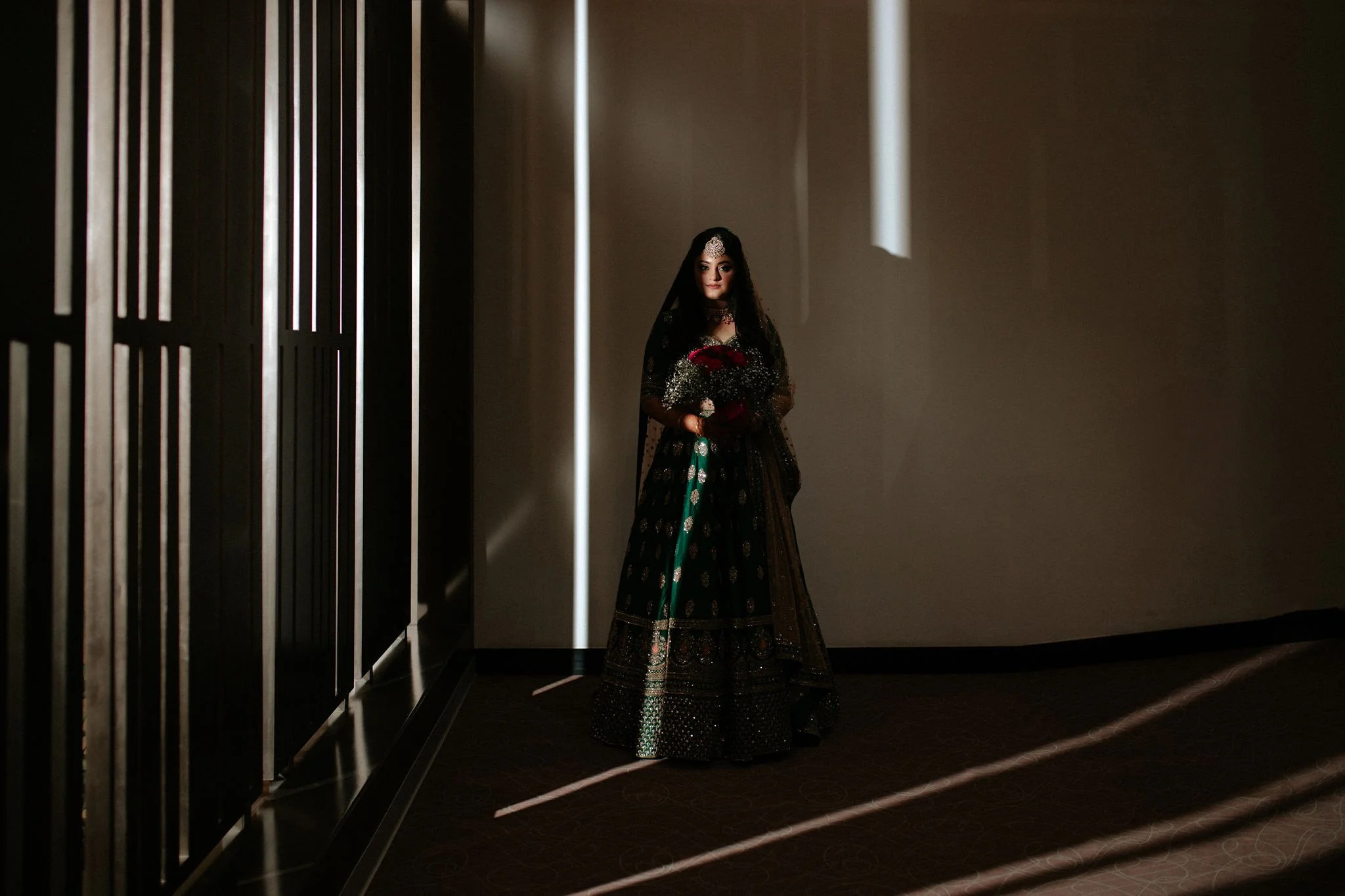 A woman dressed in elegant traditional South Asian attire, standing in a dimly lit room with shadows and light beams crossing the room.