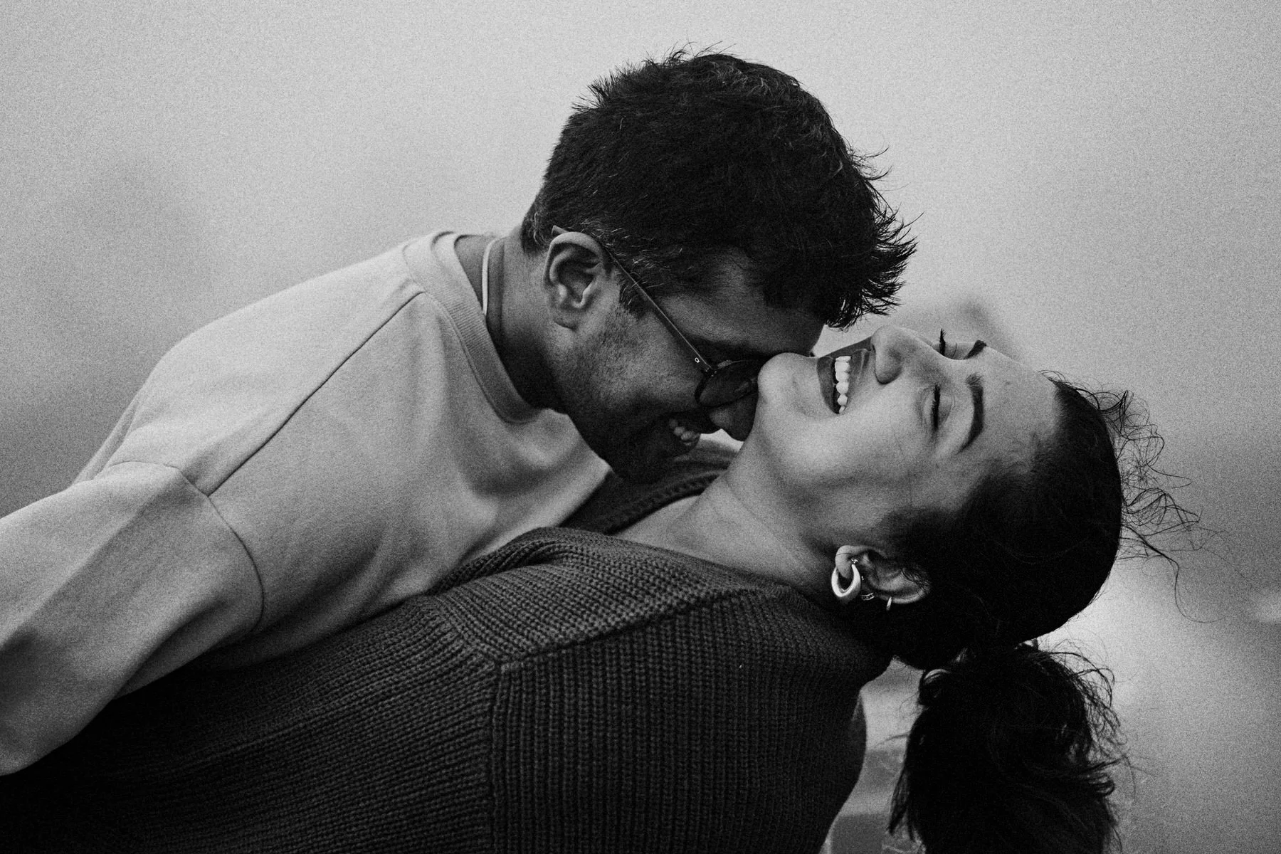 A black and white photo of a man and woman smiling and leaning close to each other, with the man kissing the woman's forehead. The woman has earrings and long hair tied back.