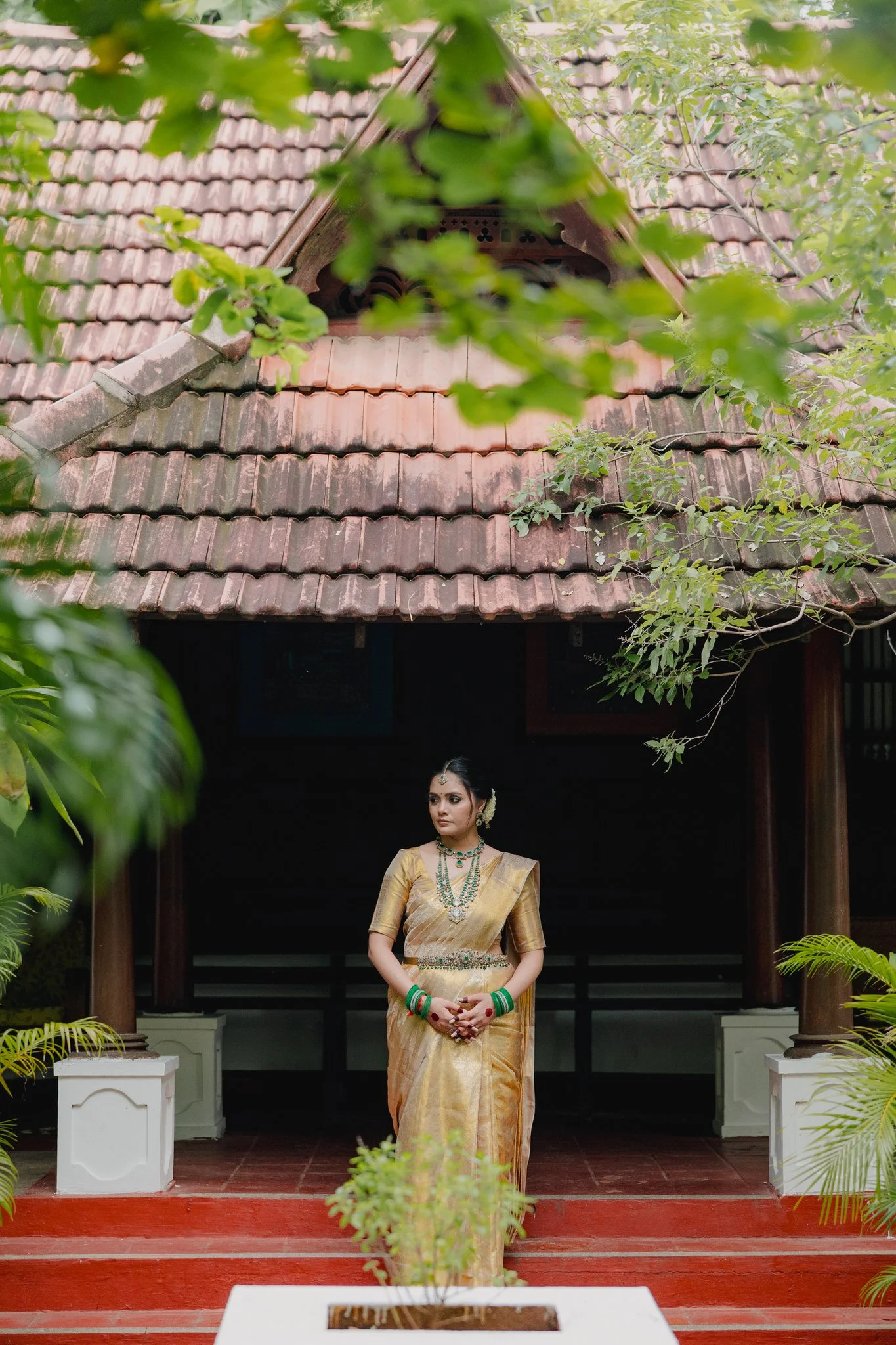 A woman dressed in traditional Indian attire standing on red steps outside a wooden building with a tiled roof, surrounded by green foliage.