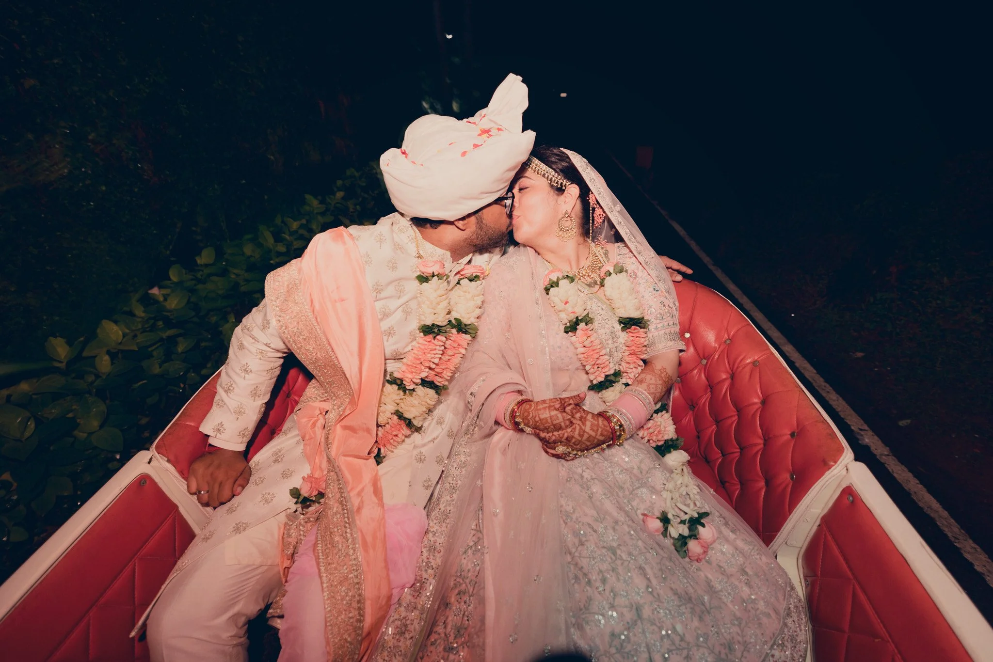 A newlywed couple in traditional Indian wedding attire sharing a kiss while sitting in a decorated cart at night.