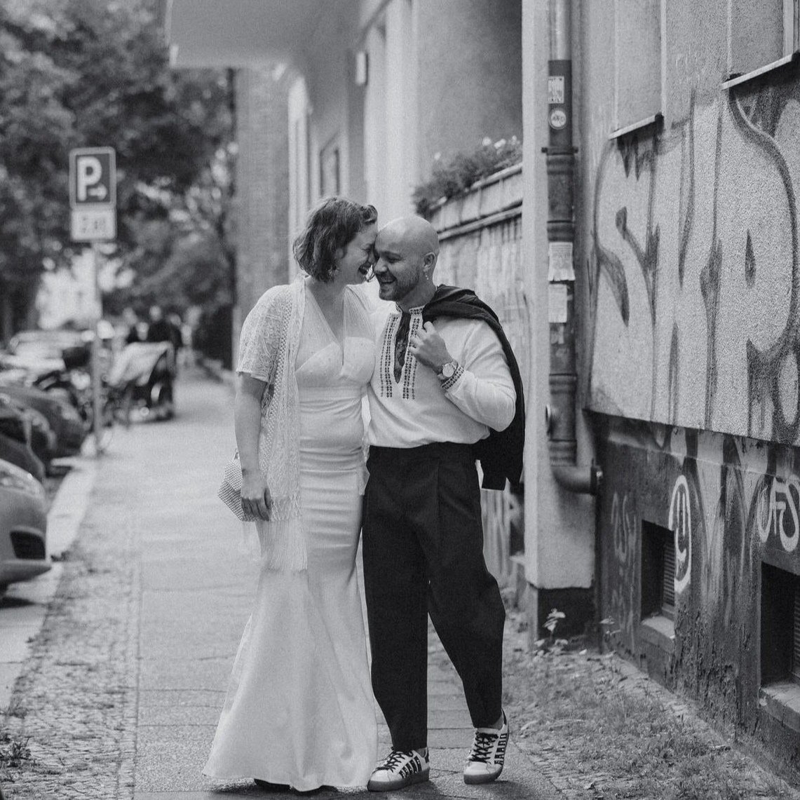 A couple in wedding attire sharing a joyful moment outside on a city sidewalk, with cars parked nearby and graffiti on the wall.