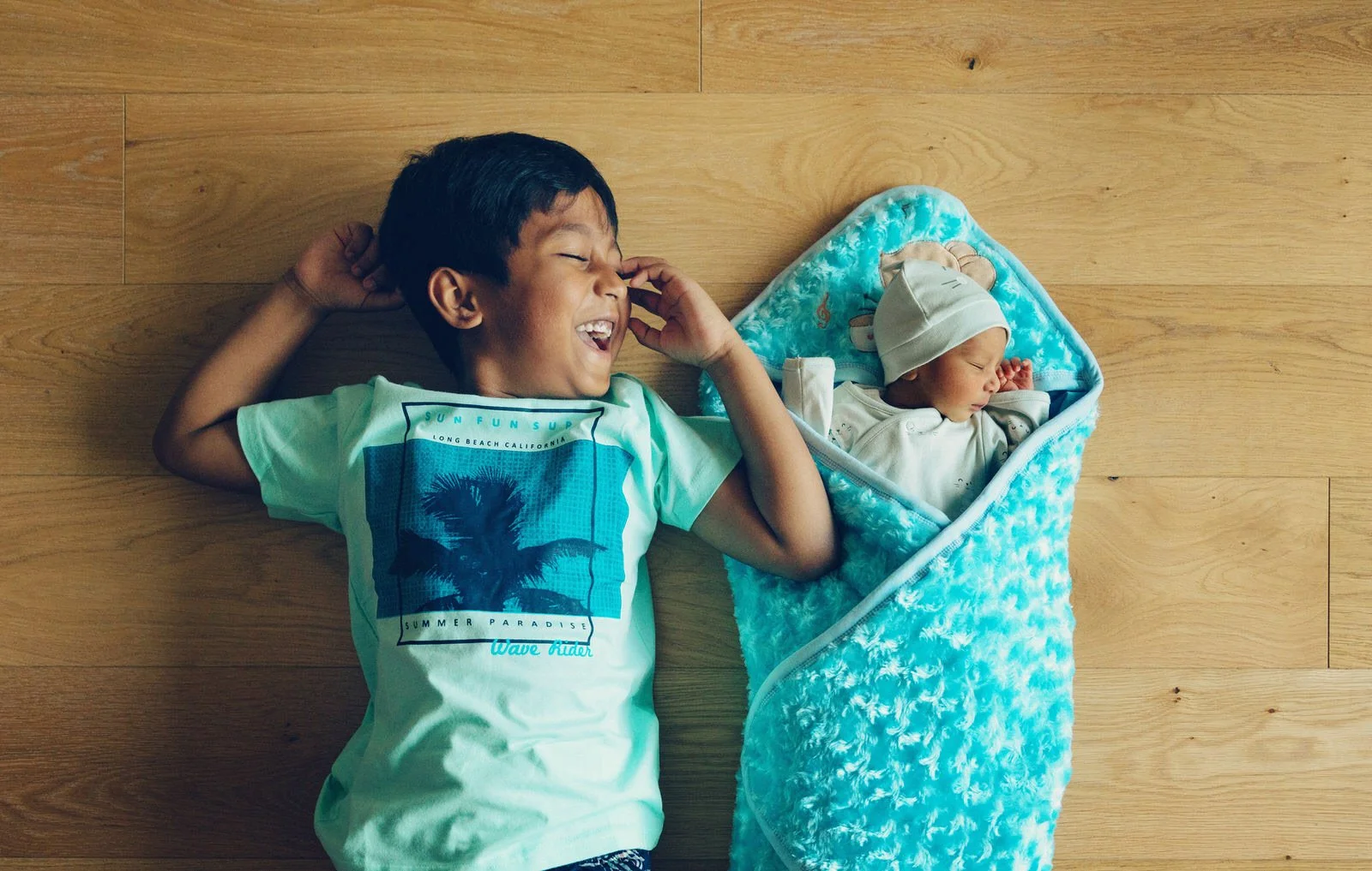 A young boy with a happy expression lying on a wooden floor next to a sleeping baby in a blue sleeping bag.