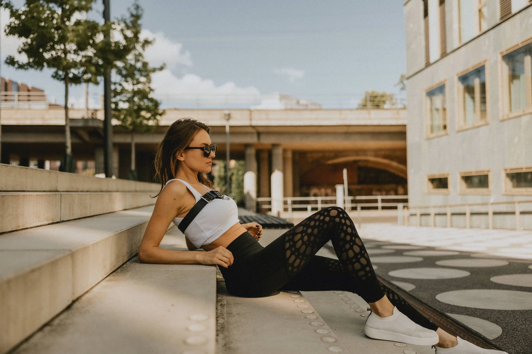 Woman sitting on concrete steps outdoors, wearing sunglasses, a white crop top, black leggings with circular cutouts, and white sneakers, with city buildings and overpass in background.