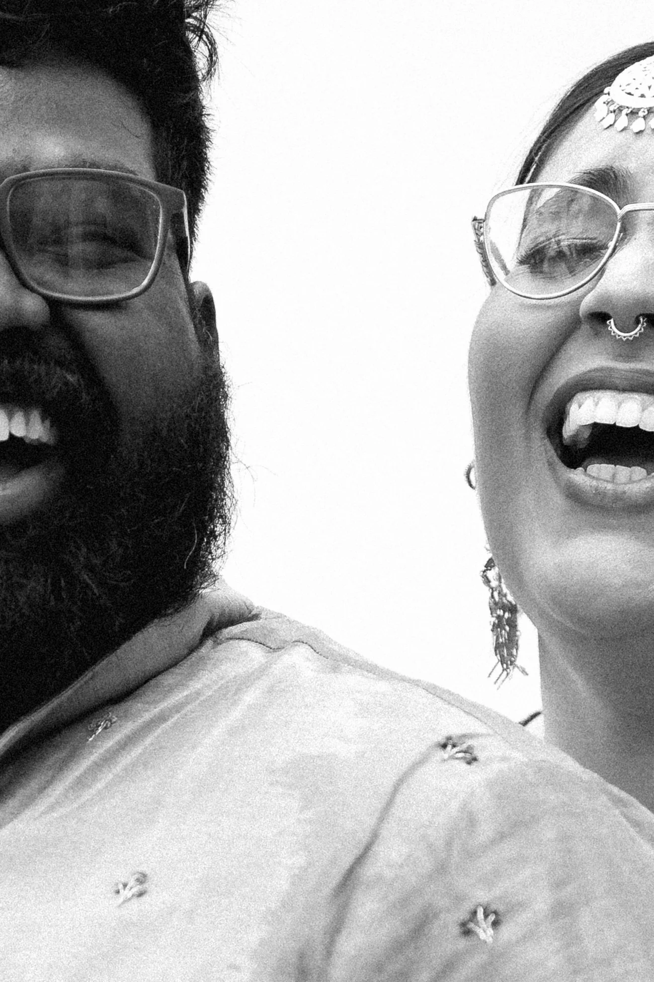 A black-and-white photo of two people smiling and laughing, wearing glasses with one person wearing a nose ring and earrings, and the other person with a beard and glasses.