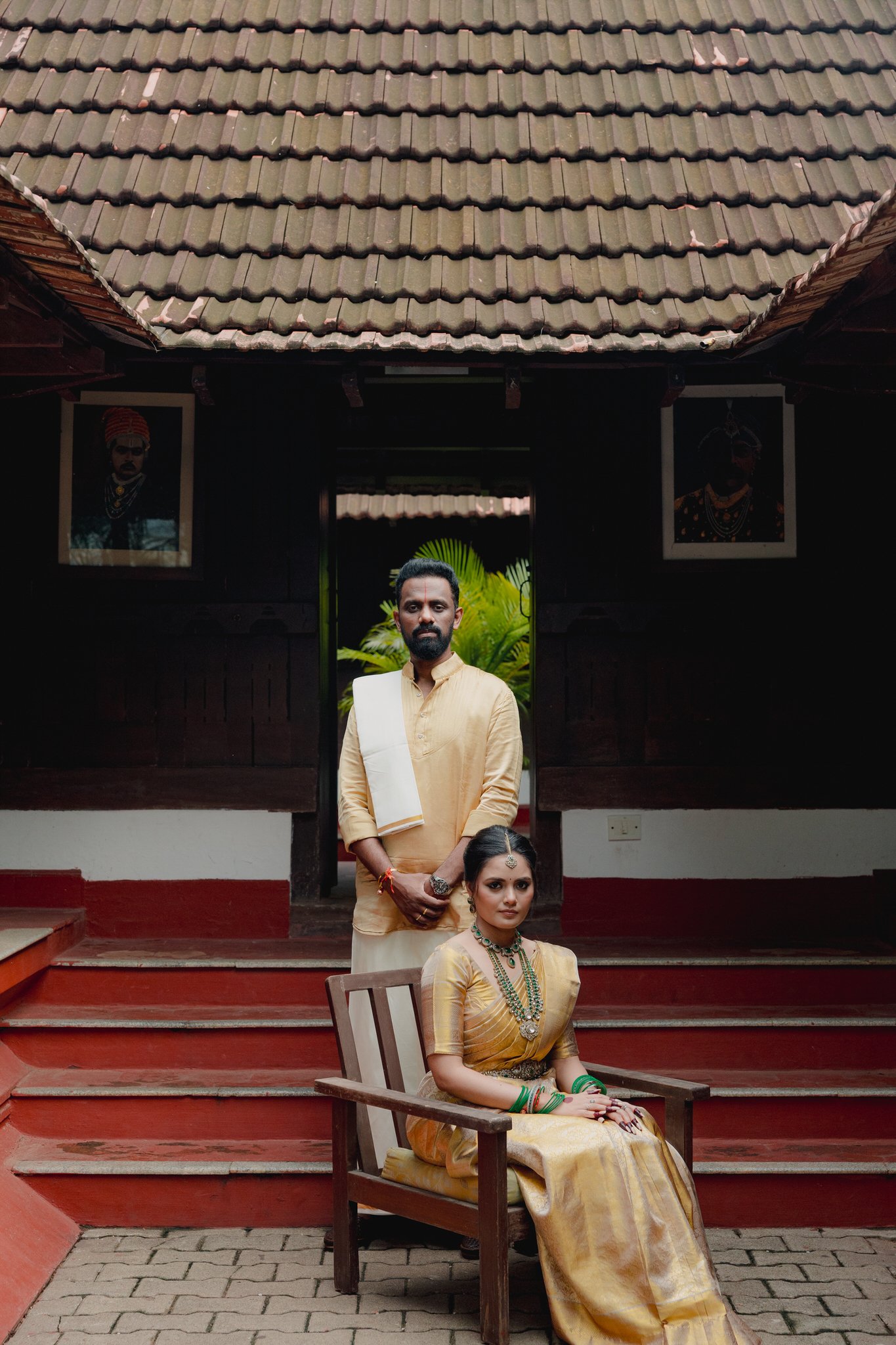 A man standing behind a seated woman, both dressed in traditional Indian attire, in front of a dark wooden house with tiled roof and framed portraits on the walls.